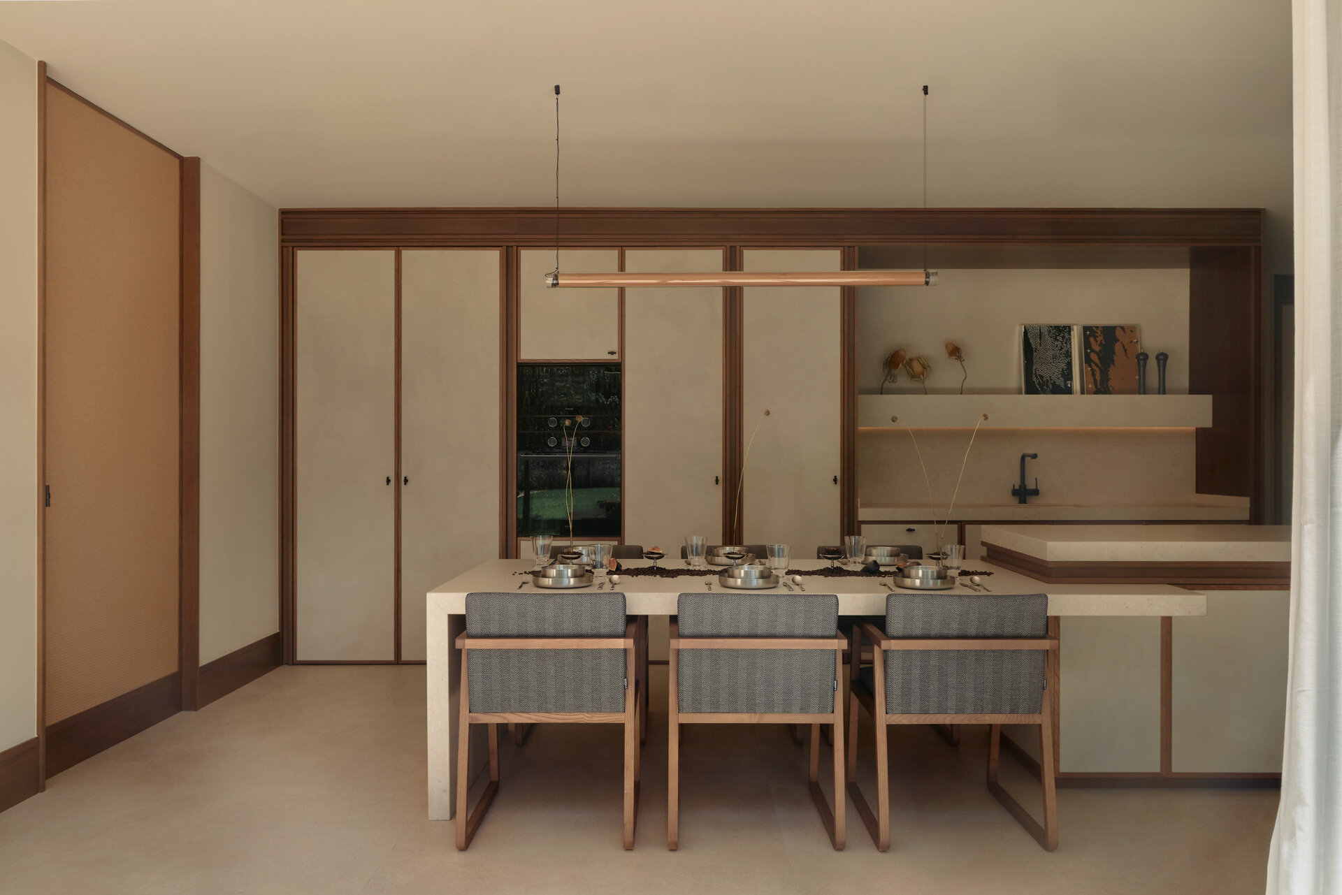 Discover a minimalist contemporary kitchen featuring microcement cabinets, timber detailing, integrated appliances, pale countertops, and an adjoining dining table with upholstered chairs.