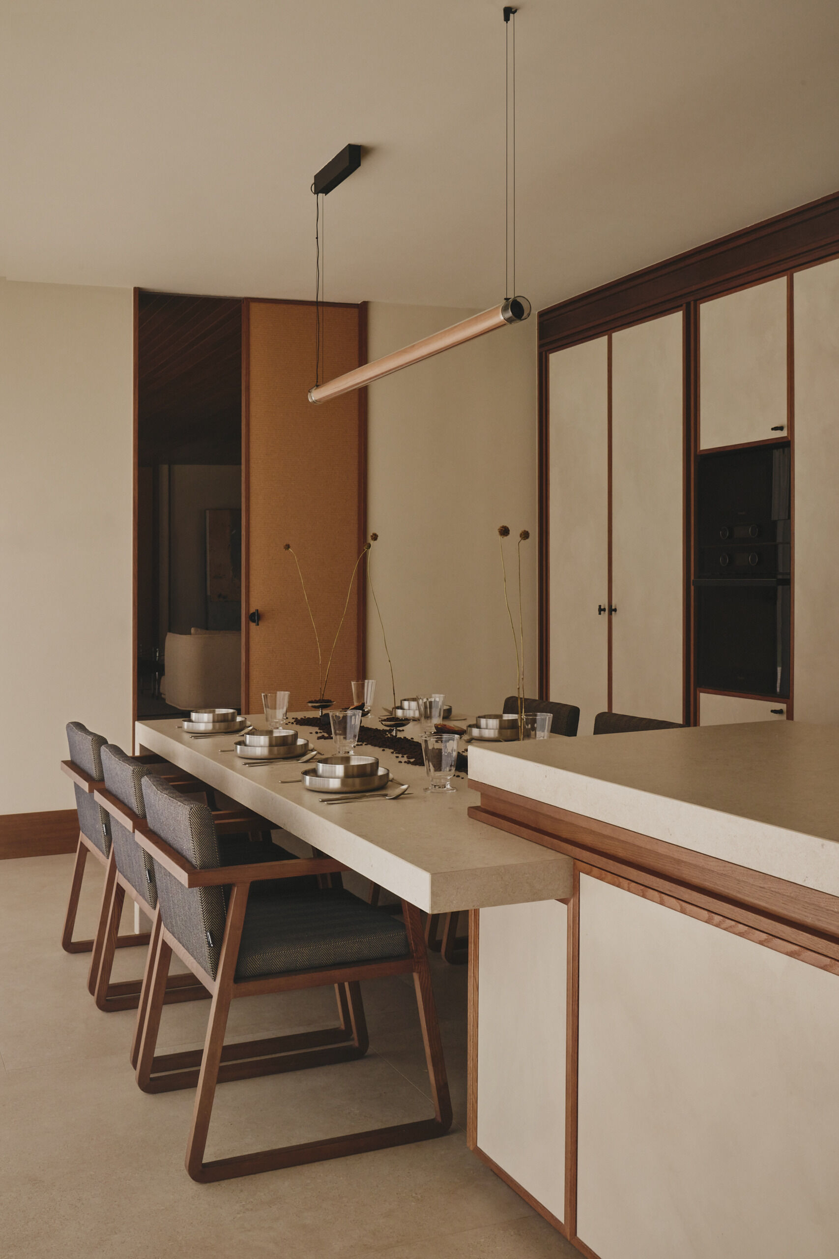 Discover a minimalist contemporary kitchen featuring microcement cabinets, timber detailing, integrated appliances, pale countertops, and an adjoining dining table with upholstered chairs.