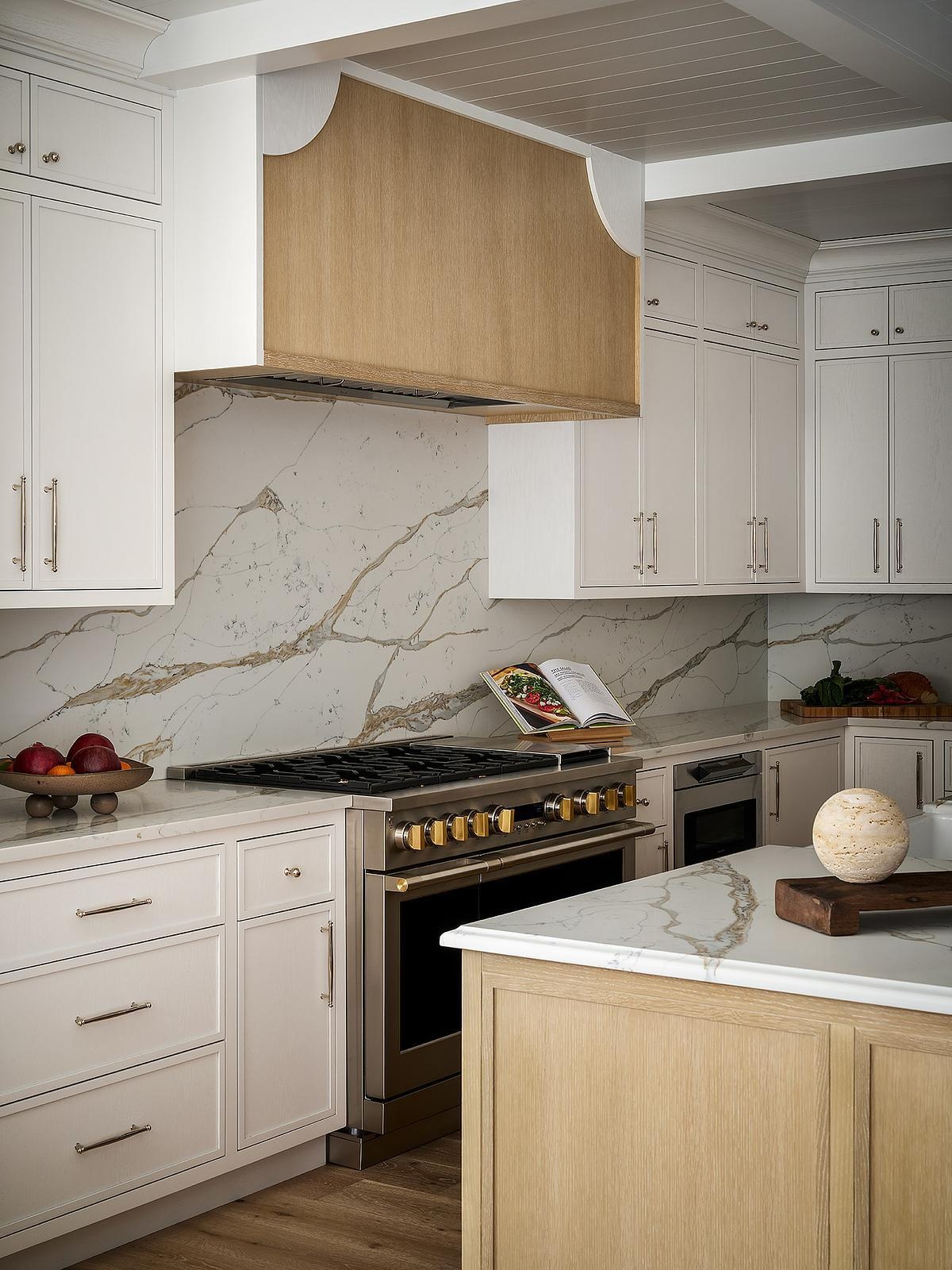 A traditional kitchen with white cabinetry, a marble-look backsplash with gold veining, a stainless steel range, and a custom wood range hood featuring curved corner details.