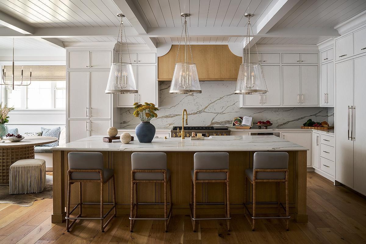 A contemporary white kitchen with a large natural wood island, woven barstools, three glass pendant lights, stone countertops, and a decorative wood range hood beneath a white beamed ceiling.