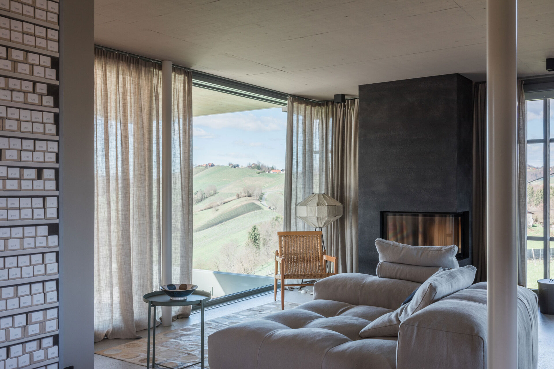 A contemporary living room in a renovated Austrian farmhouse features a large sectional sofa, dark fireplace, woven armchair, linen curtains, and panoramic countryside views.