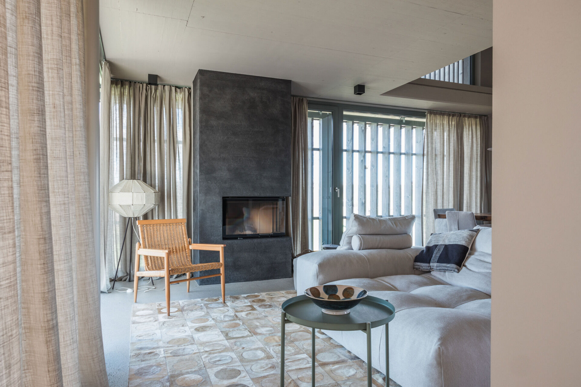 A contemporary living room in a renovated Austrian farmhouse features a large sectional sofa, dark fireplace, woven armchair, linen curtains, and panoramic countryside views.