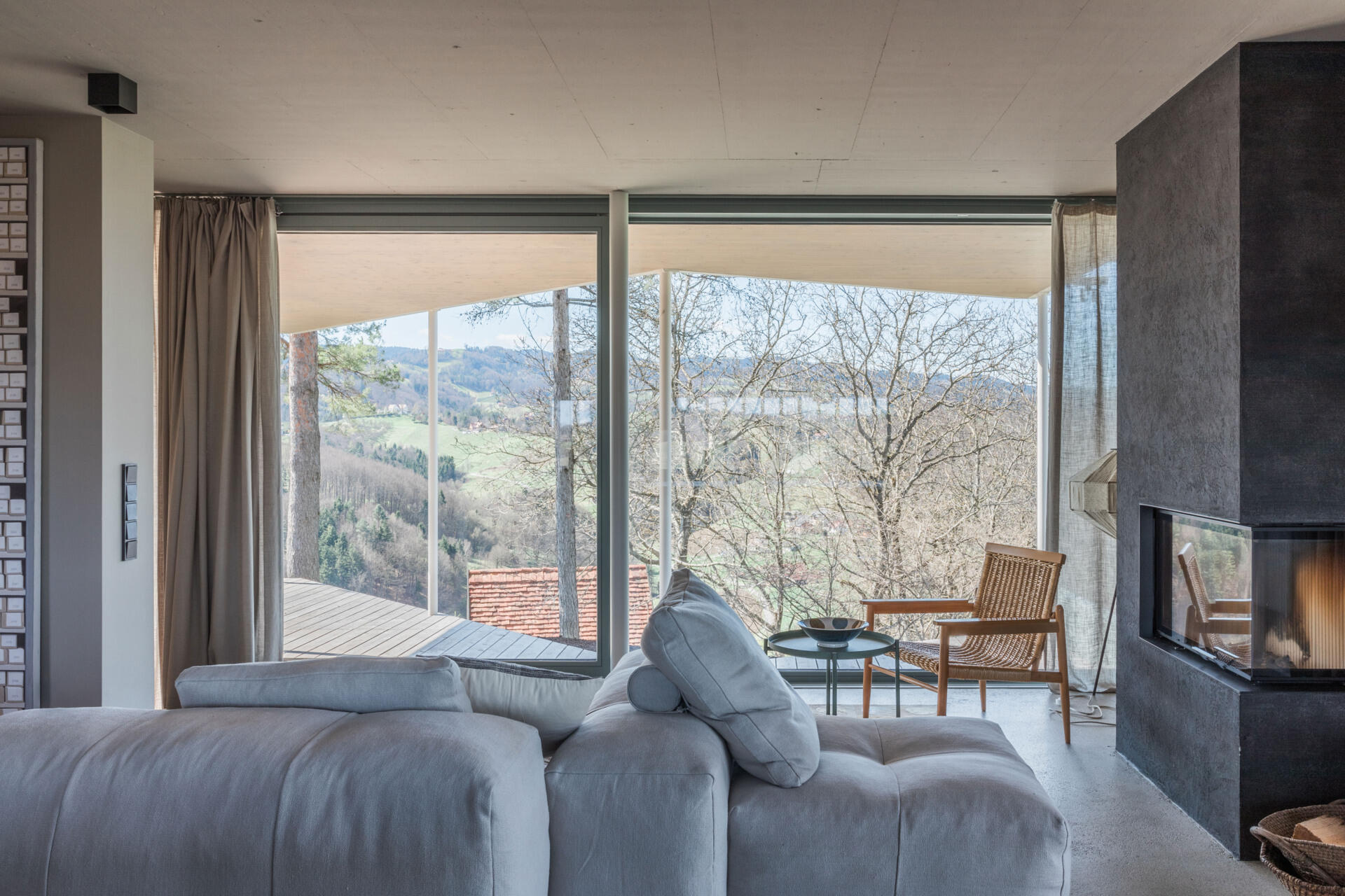A contemporary living room in a renovated Austrian farmhouse features a large sectional sofa, dark fireplace, woven armchair, linen curtains, and panoramic countryside views.