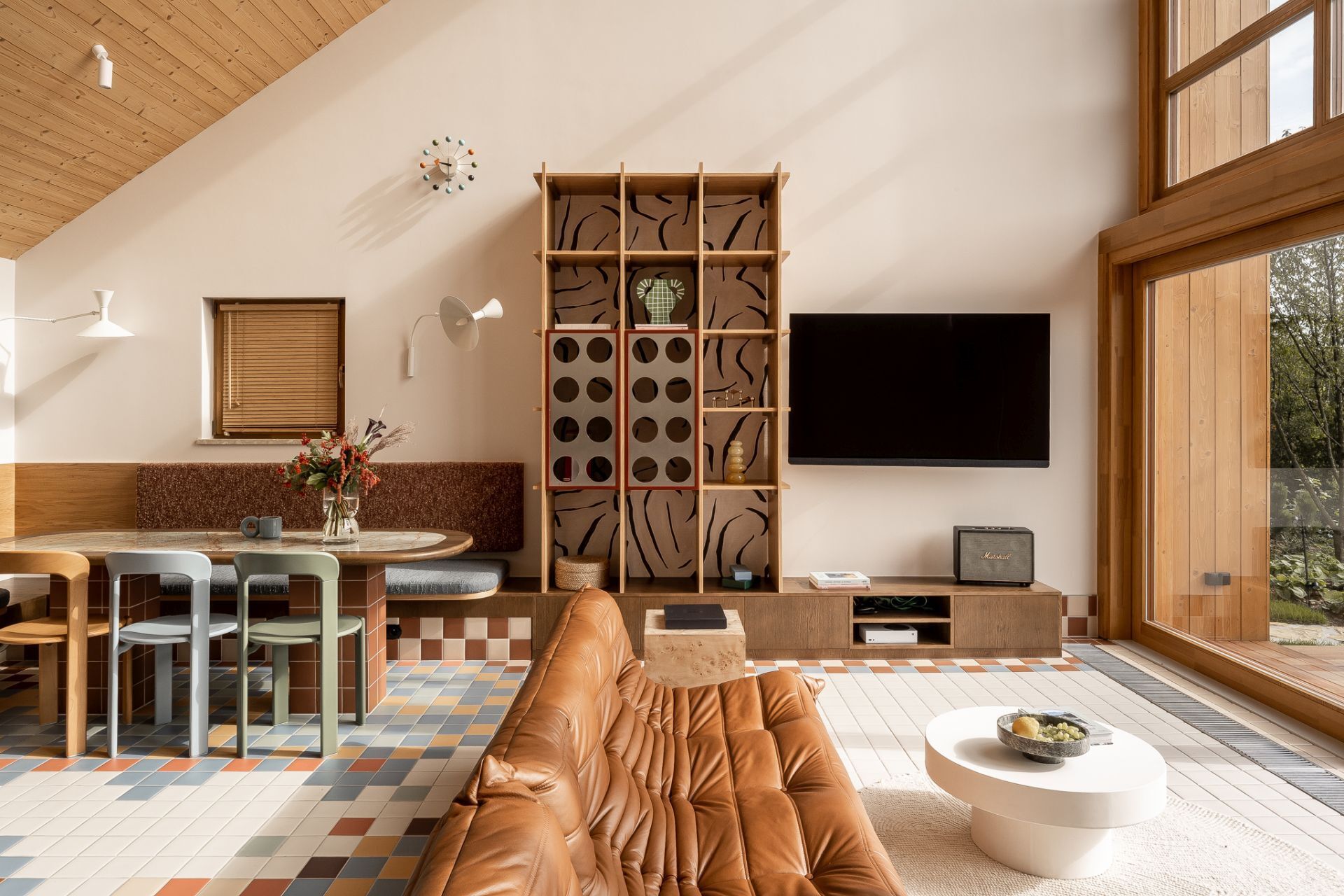 Tour a contemporary living room in Poland featuring a caramel Togo sofa, Isamu Noguchi Akari lamp, Le Corbusier wall lights, custom bookcase, and floor-to-ceiling windows overlooking a lake.