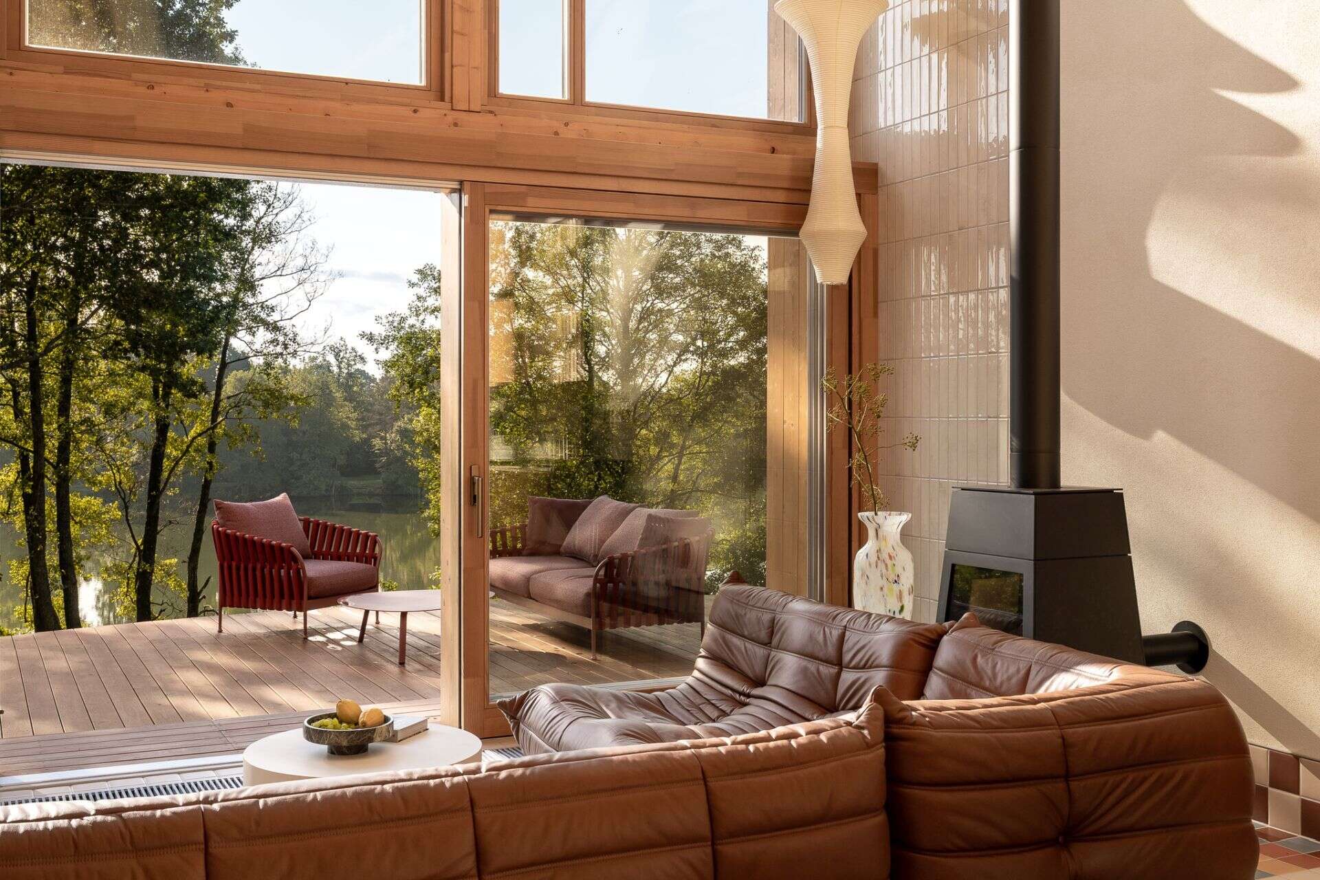 Tour a contemporary living room in Poland featuring a caramel Togo sofa, Isamu Noguchi Akari lamp, Le Corbusier wall lights, custom bookcase, and floor-to-ceiling windows overlooking a lake.