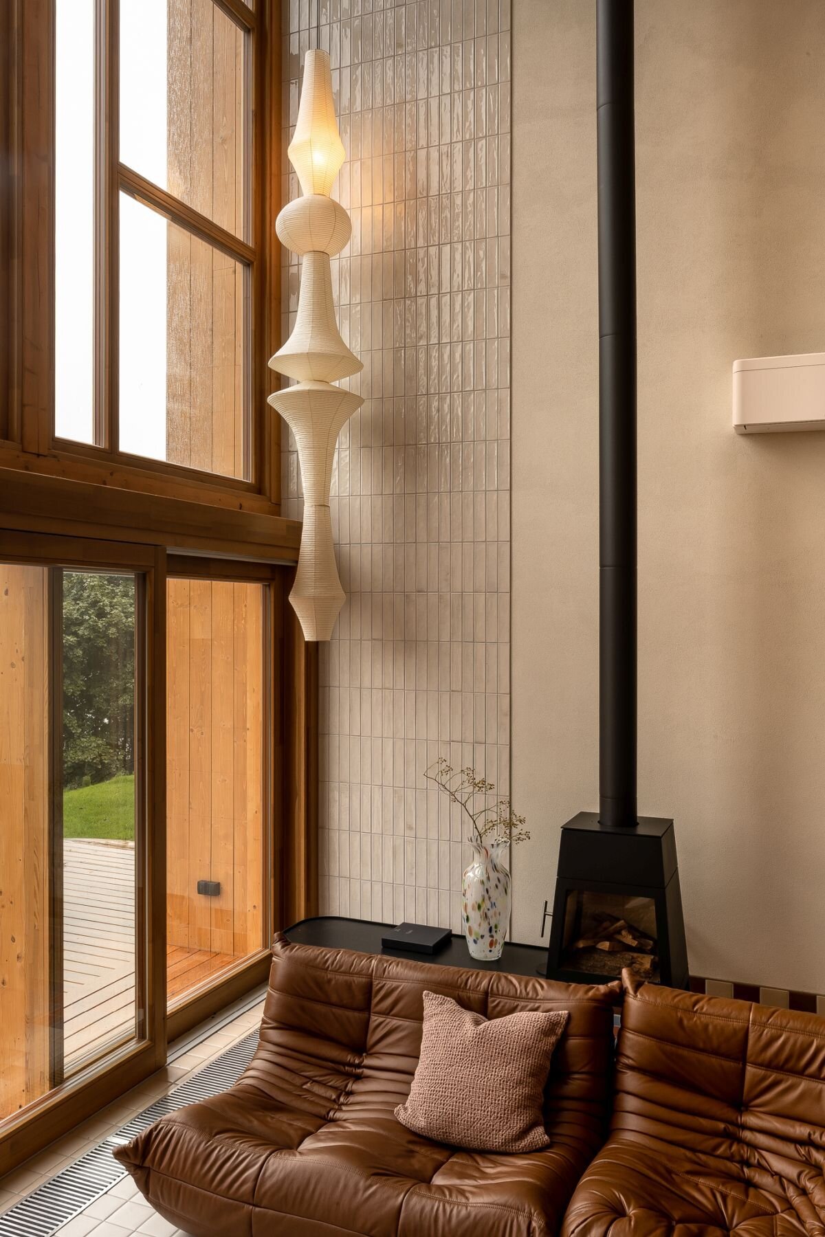 Tour a contemporary living room in Poland featuring a caramel Togo sofa, Isamu Noguchi Akari lamp, Le Corbusier wall lights, custom bookcase, and floor-to-ceiling windows overlooking a lake.