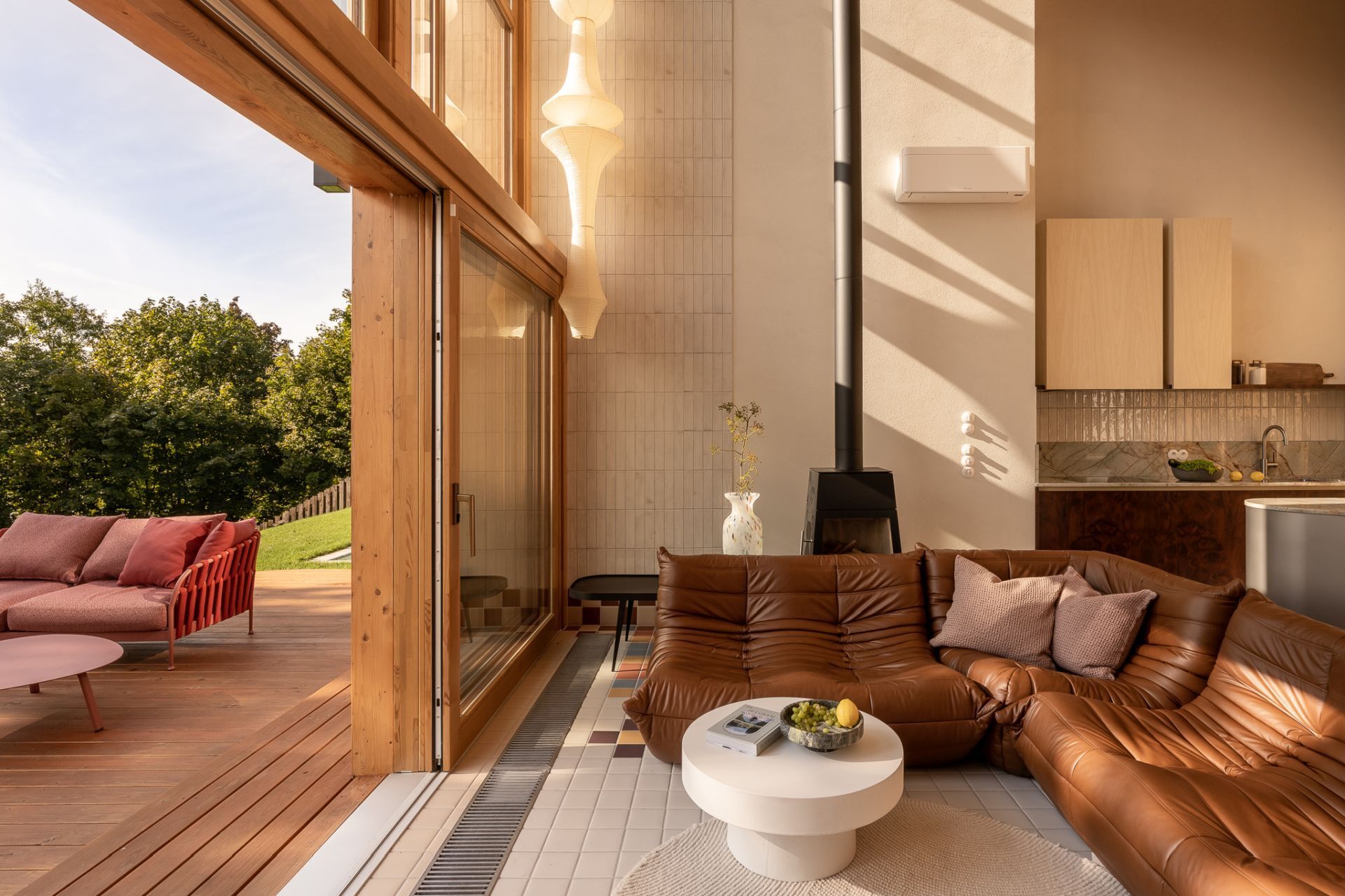 Tour a contemporary living room in Poland featuring a caramel Togo sofa, Isamu Noguchi Akari lamp, Le Corbusier wall lights, custom bookcase, and floor-to-ceiling windows overlooking a lake.