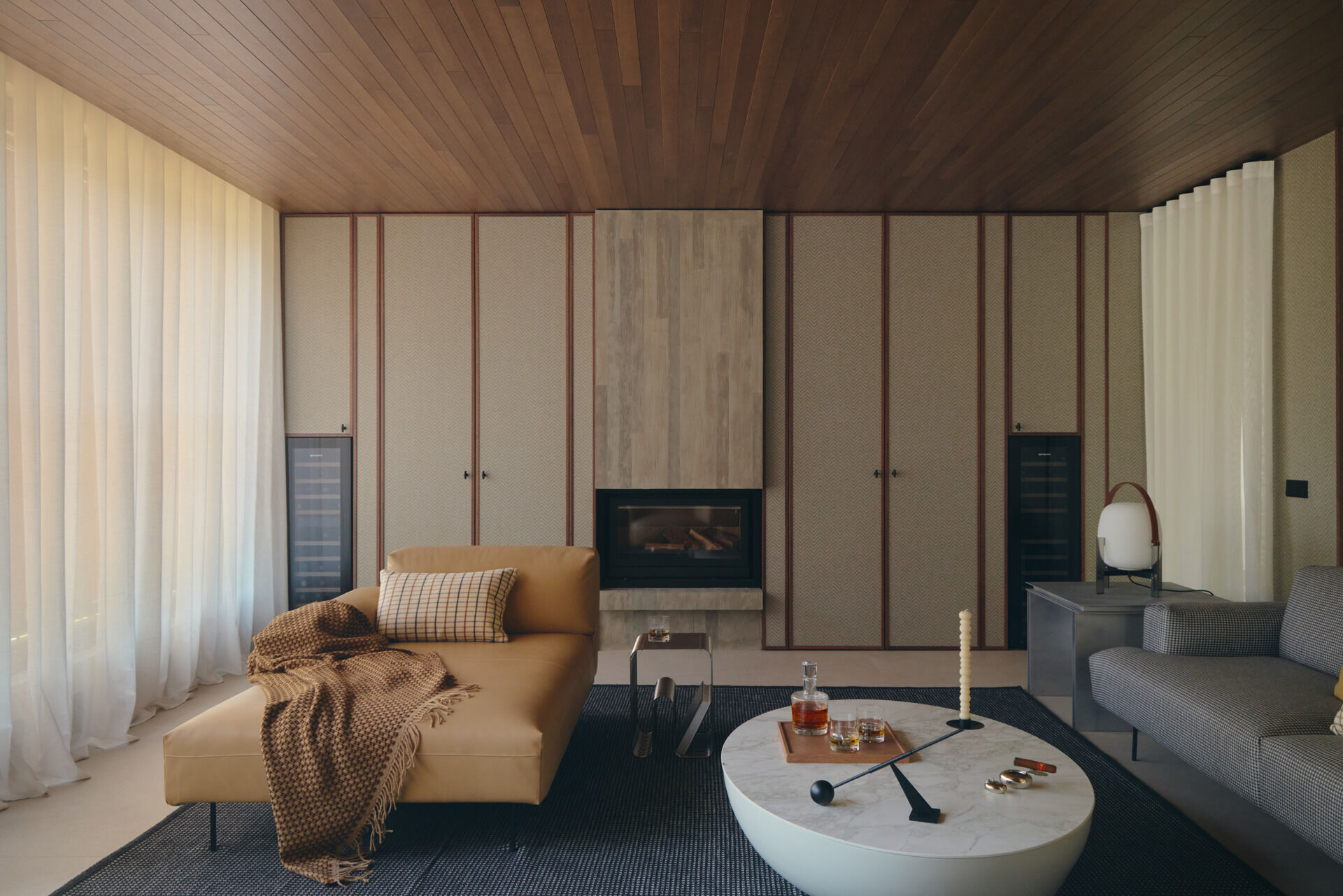 Step inside a contemporary cigar lounge with herringbone wallpaper, a concrete fireplace, caramel-colored lounge chairs, a marble coffee table, and designer lighting.