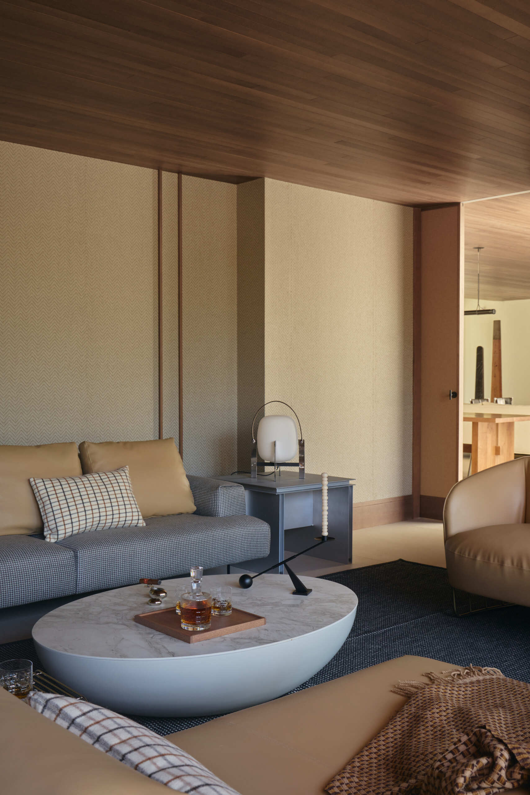 Step inside a contemporary cigar lounge with herringbone wallpaper, a concrete fireplace, caramel-colored lounge chairs, a marble coffee table, and designer lighting.