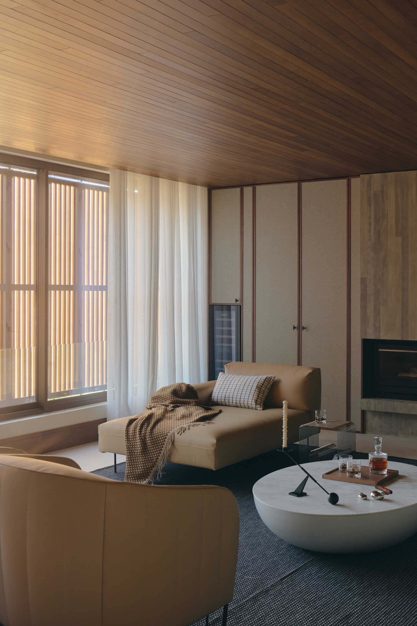 Step inside a contemporary cigar lounge with herringbone wallpaper, a concrete fireplace, caramel-colored lounge chairs, a marble coffee table, and designer lighting.