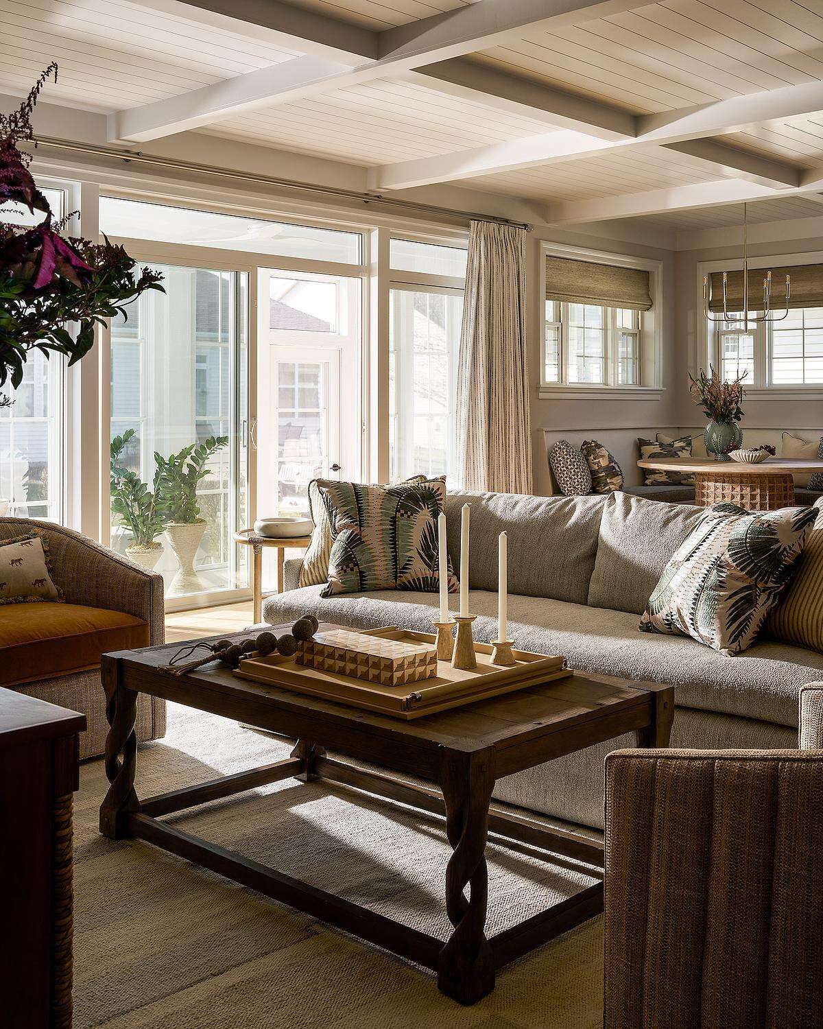 A coastal-inspired living room featuring neutral upholstered sofas, botanical patterned pillows, a dark wood coffee table, white ceiling beams, and large glass doors that fill the room with natural light.