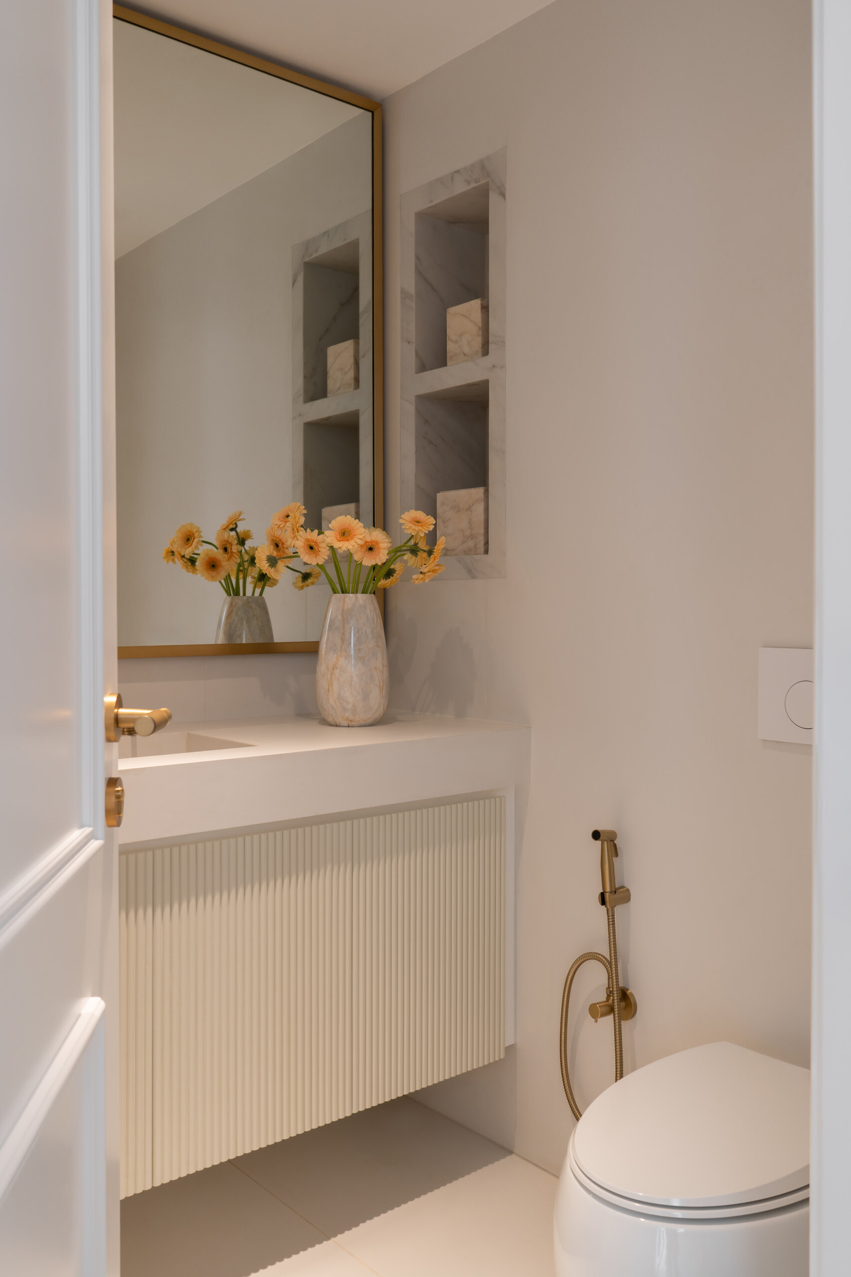 This elegant powder room in The White Arcade by D’Ora Tokai Designs features a floating ribbed vanity, white countertop, oversized brass-framed mirror, built-in marble niches, and warm brass bathroom fixtures. Minimalist luxury bathroom inspiration from Dubai.