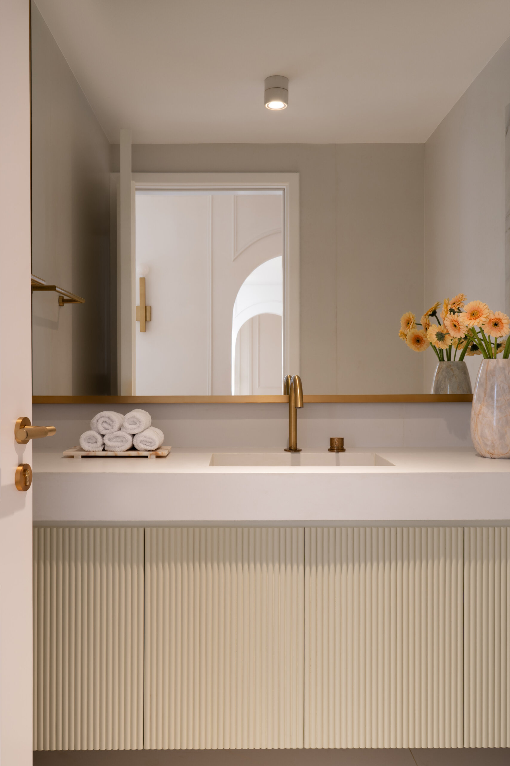 This elegant powder room in The White Arcade by D’Ora Tokai Designs features a floating ribbed vanity, white countertop, oversized brass-framed mirror, built-in marble niches, and warm brass bathroom fixtures. Minimalist luxury bathroom inspiration from Dubai.