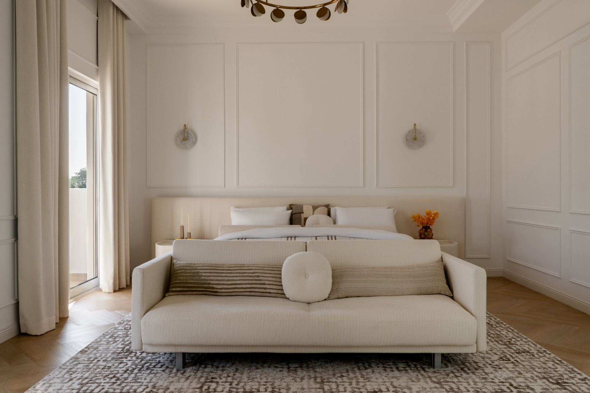 Inside the luxurious primary bedroom of The White Arcade in Dubai, designed by D’Ora Tokai Designs. The spacious suite features a bespoke upholstered bed, cream sofa, sculptural lounge chair, brass chandelier, and soft neutral furnishings with access to a private balcony.