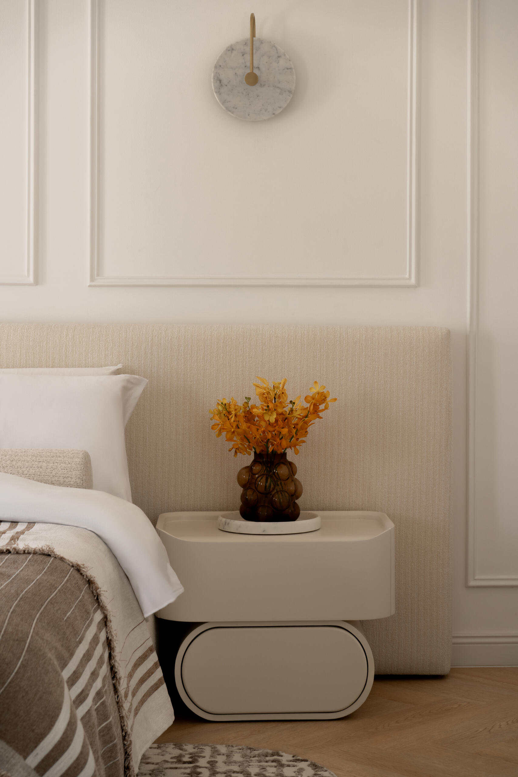Inside the luxurious primary bedroom of The White Arcade in Dubai, designed by D’Ora Tokai Designs. The spacious suite features a bespoke upholstered bed, cream sofa, sculptural lounge chair, brass chandelier, and soft neutral furnishings with access to a private balcony.