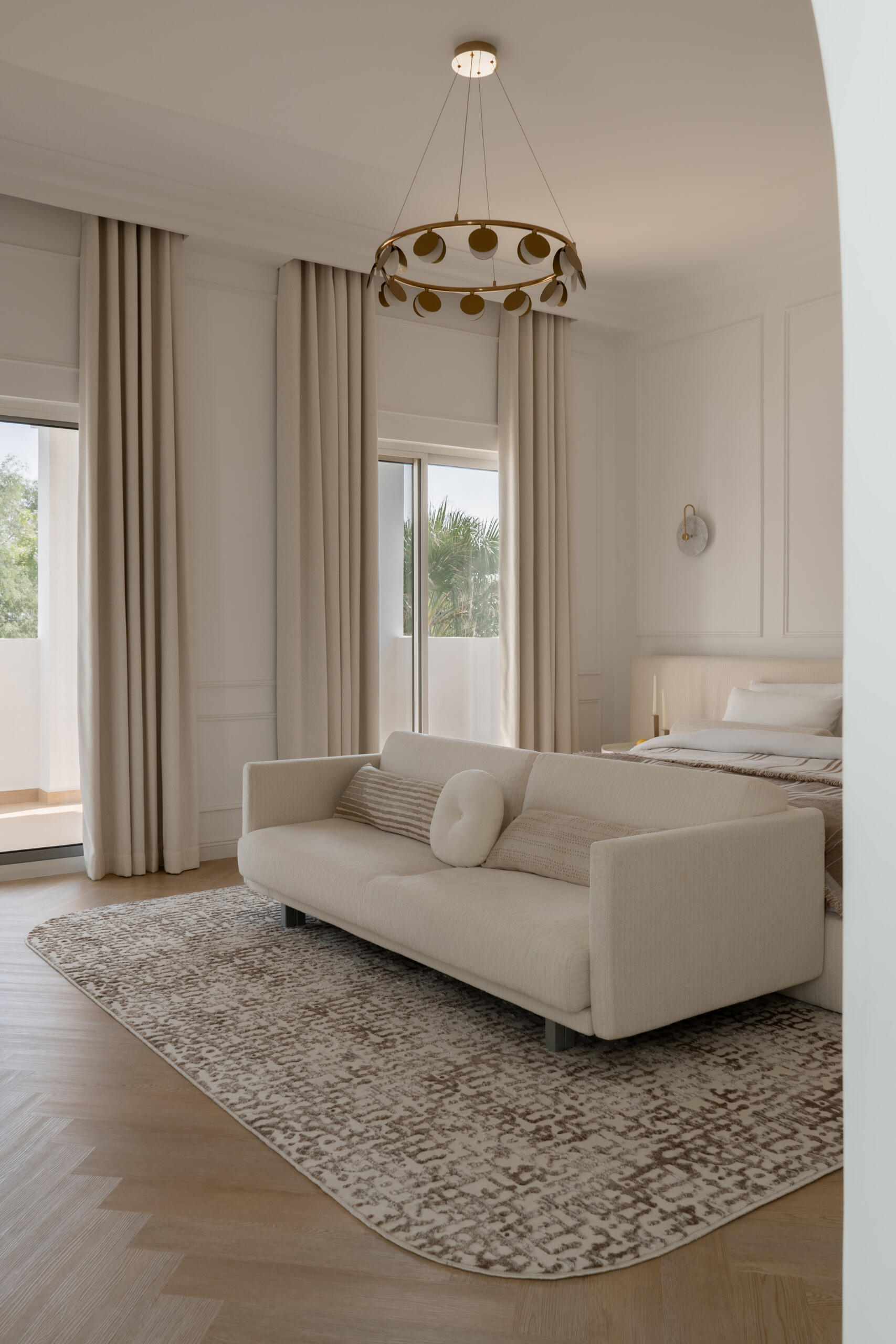Inside the luxurious primary bedroom of The White Arcade in Dubai, designed by D’Ora Tokai Designs. The spacious suite features a bespoke upholstered bed, cream sofa, sculptural lounge chair, brass chandelier, and soft neutral furnishings with access to a private balcony.