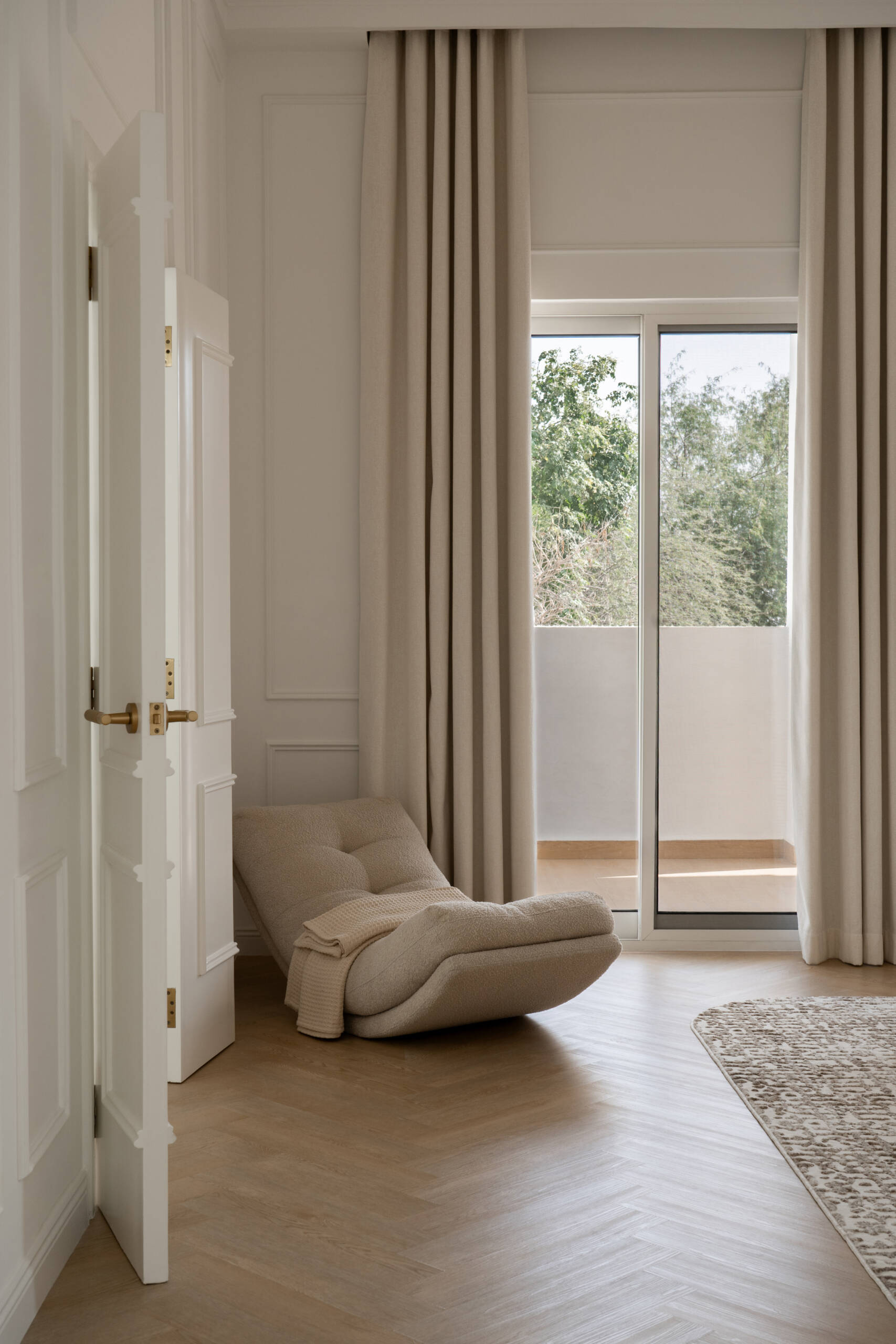 Inside the luxurious primary bedroom of The White Arcade in Dubai, designed by D’Ora Tokai Designs. The spacious suite features a bespoke upholstered bed, cream sofa, sculptural lounge chair, brass chandelier, and soft neutral furnishings with access to a private balcony.