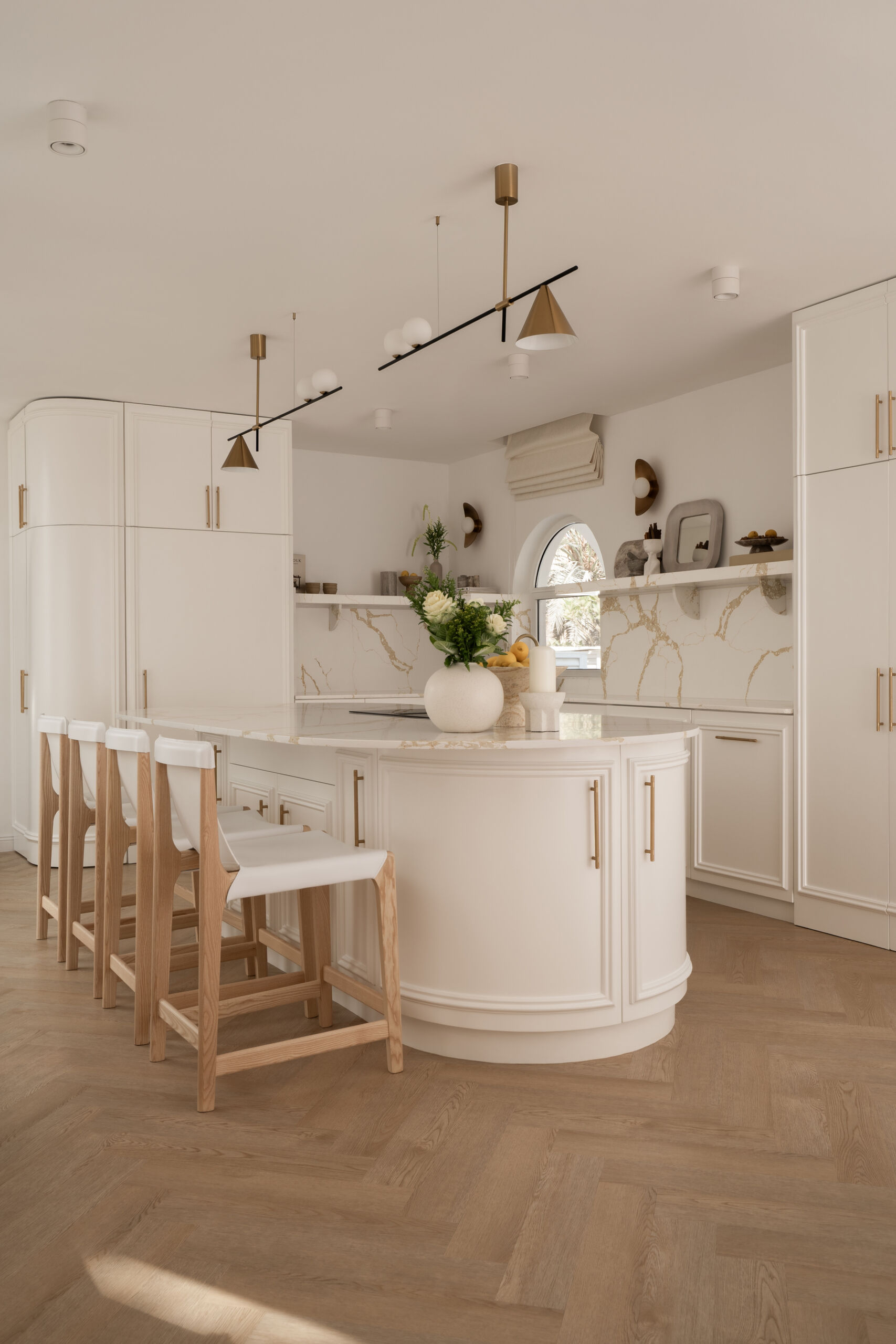 Explore this luxurious white kitchen by D’Ora Tokai Designs in Dubai. Featuring a large curved kitchen island, white stone countertops with gold veining, brass cabinet handles, arched windows, and contemporary pendant lighting, The White Arcade offers beautiful inspiration for an elegant modern kitchen.