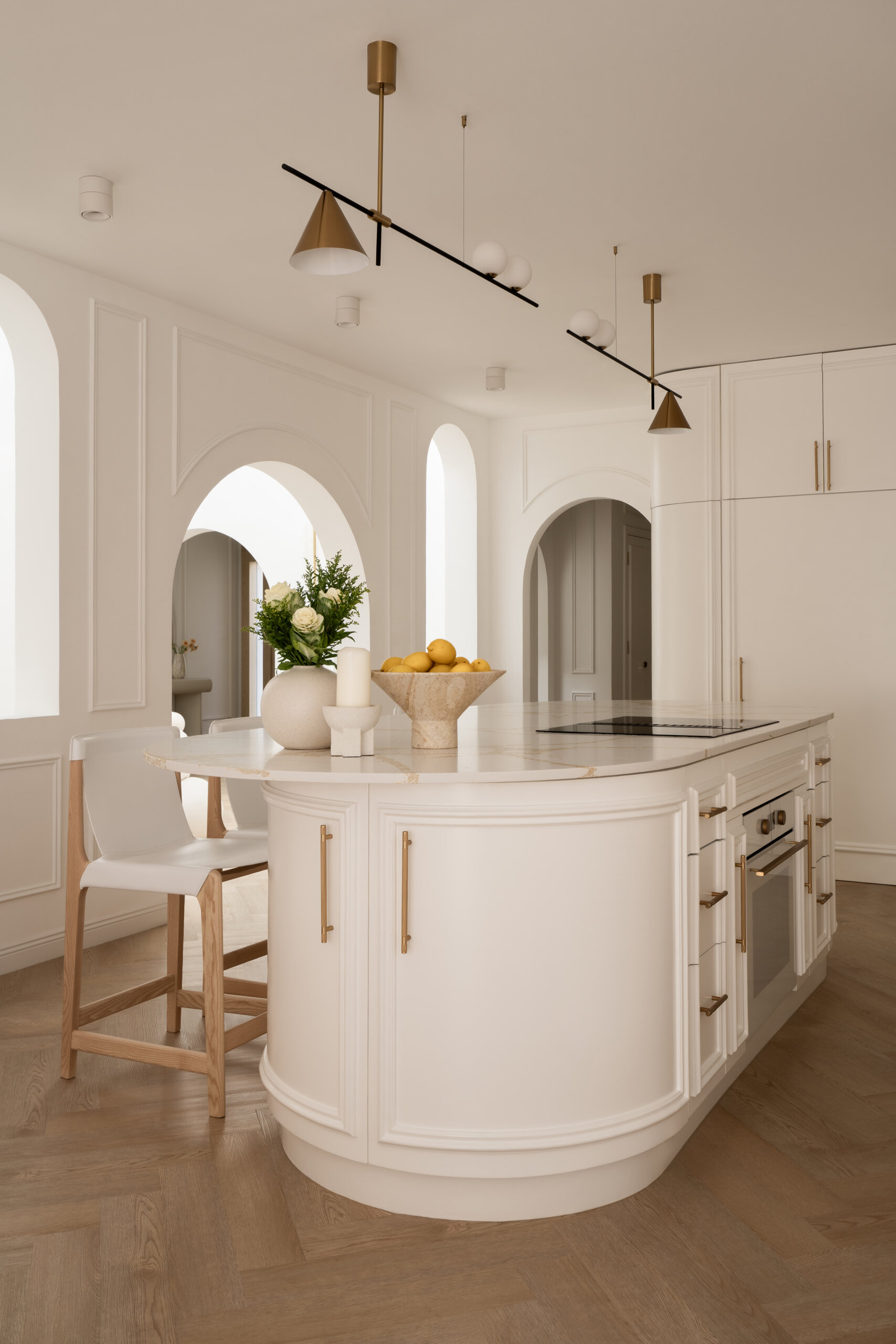 Explore this luxurious white kitchen by D’Ora Tokai Designs in Dubai. Featuring a large curved kitchen island, white stone countertops with gold veining, brass cabinet handles, arched windows, and contemporary pendant lighting, The White Arcade offers beautiful inspiration for an elegant modern kitchen.
