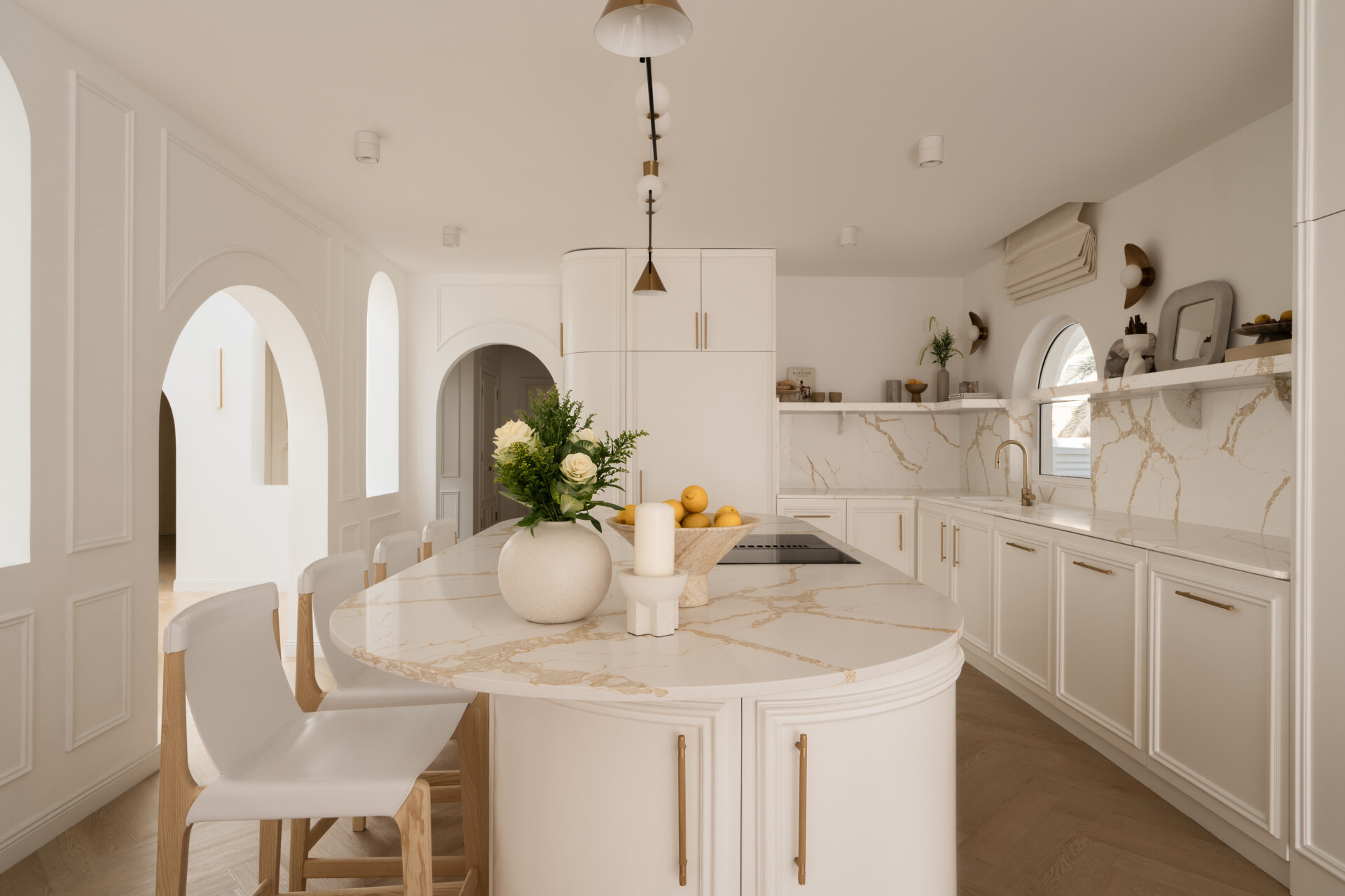 Explore this luxurious white kitchen by D’Ora Tokai Designs in Dubai. Featuring a large curved kitchen island, white stone countertops with gold veining, brass cabinet handles, arched windows, and contemporary pendant lighting, The White Arcade offers beautiful inspiration for an elegant modern kitchen.