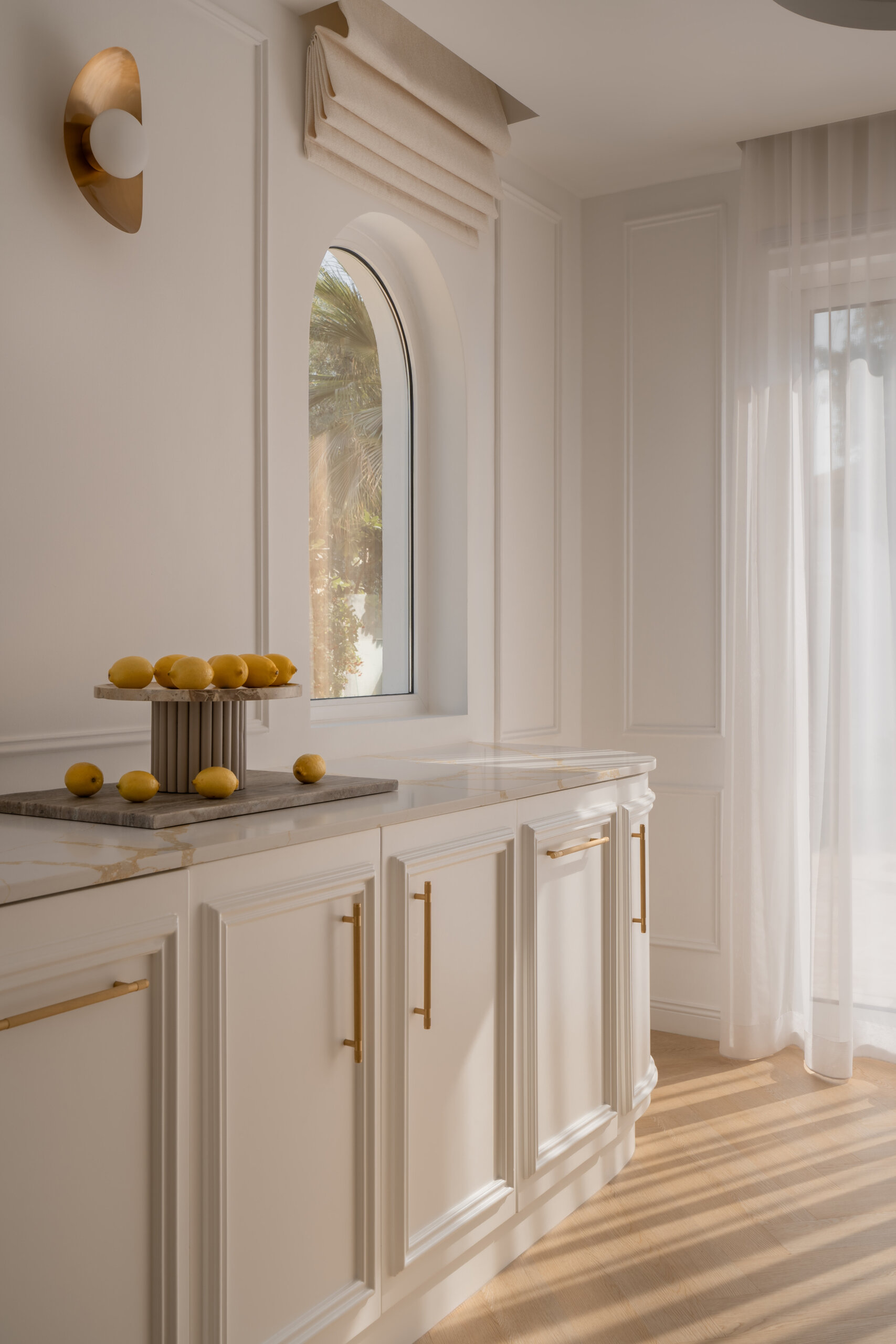 A contemporary casual dining room inside The White Arcade in Dubai, featuring sculptural white dining chairs, an oval table, a circular brass chandelier, arched windows, and elegant built-in cabinetry. Beautiful neutral dining room inspiration by D’Ora Tokai Designs.