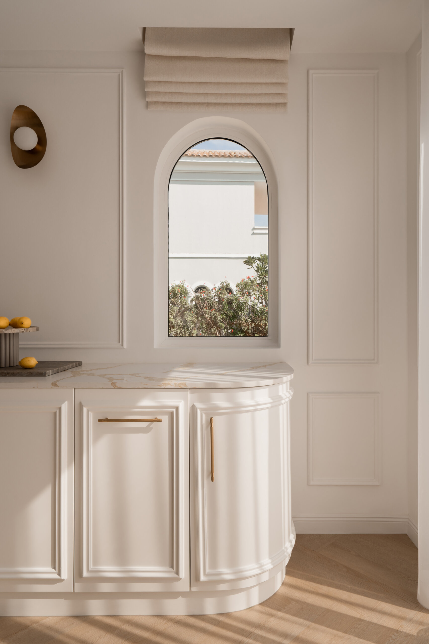 A contemporary casual dining room inside The White Arcade in Dubai, featuring sculptural white dining chairs, an oval table, a circular brass chandelier, arched windows, and elegant built-in cabinetry. Beautiful neutral dining room inspiration by D’Ora Tokai Designs.