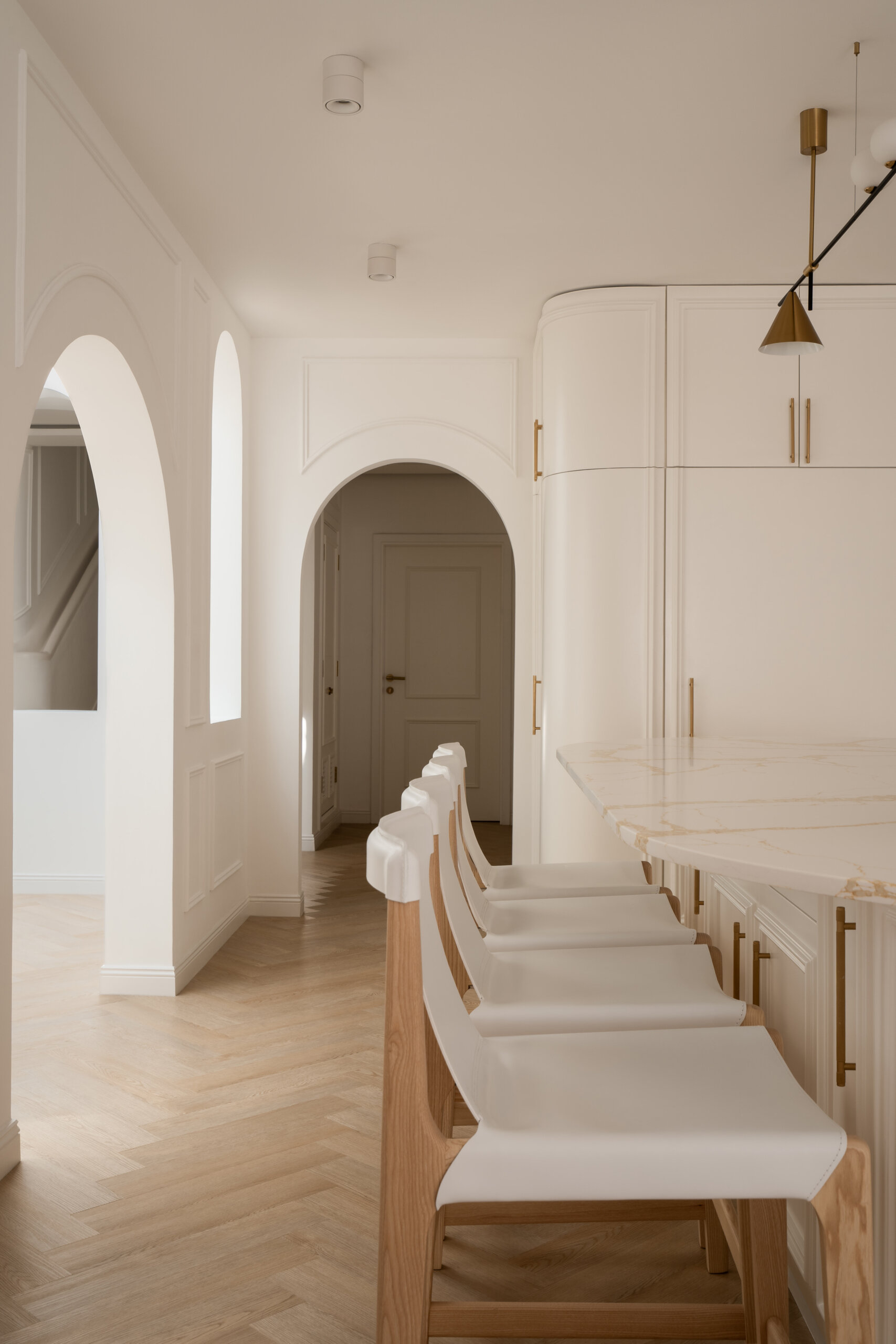 Explore this luxurious white kitchen by D’Ora Tokai Designs in Dubai. Featuring a large curved kitchen island, white stone countertops with gold veining, brass cabinet handles, arched windows, and contemporary pendant lighting, The White Arcade offers beautiful inspiration for an elegant modern kitchen.