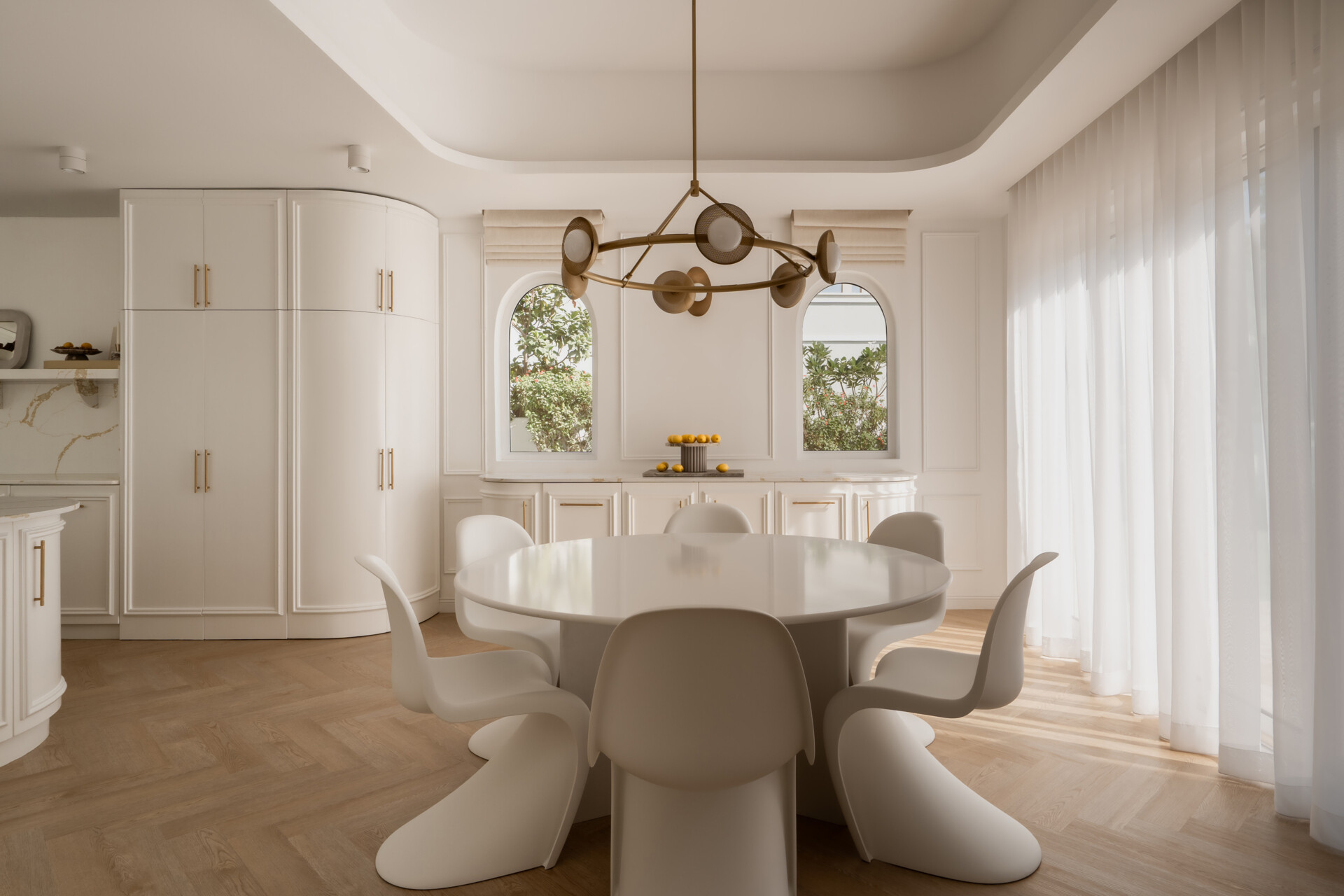 A contemporary casual dining room inside The White Arcade in Dubai, featuring sculptural white dining chairs, an oval table, a circular brass chandelier, arched windows, and elegant built-in cabinetry. Beautiful neutral dining room inspiration by D’Ora Tokai Designs.