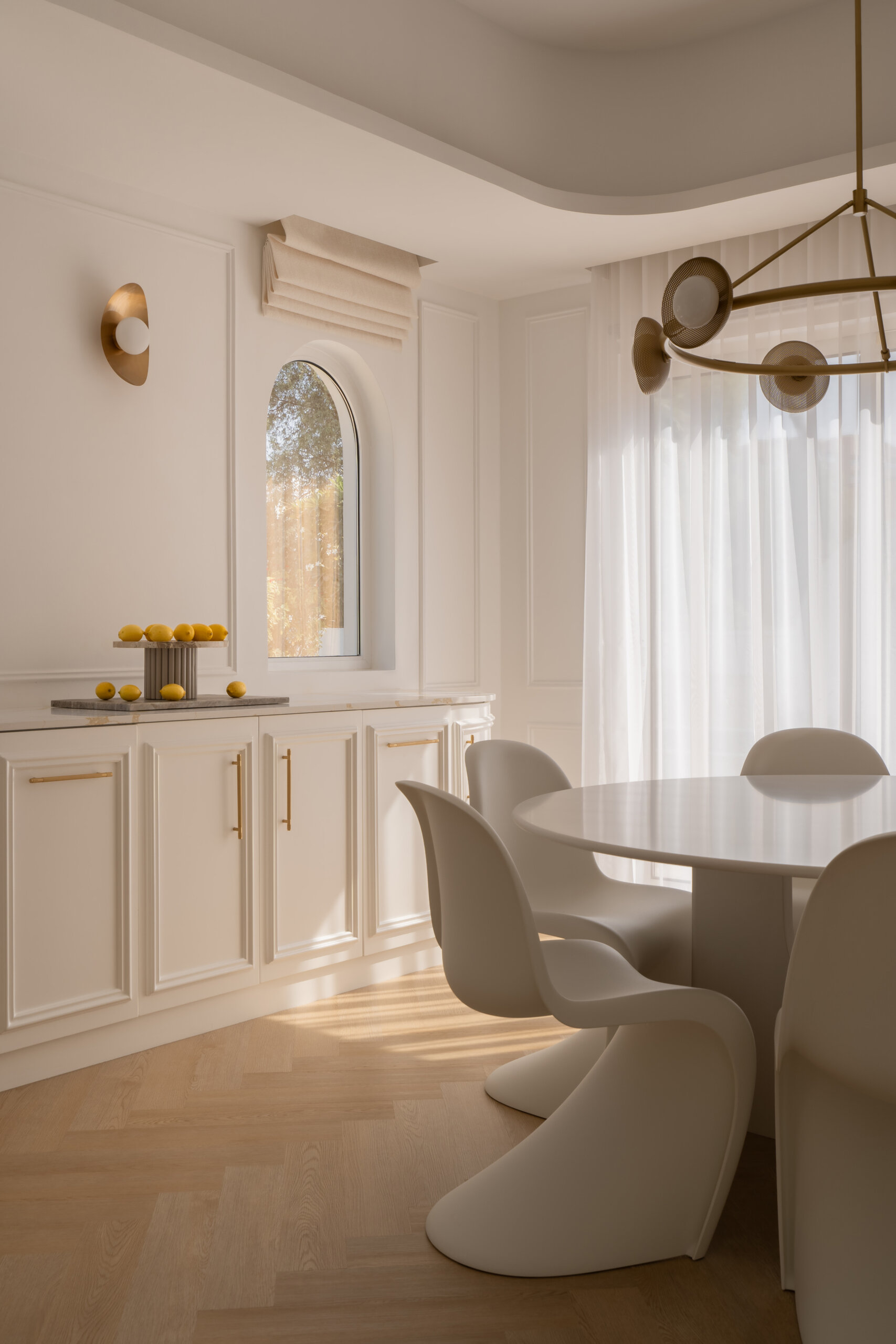 A contemporary casual dining room inside The White Arcade in Dubai, featuring sculptural white dining chairs, an oval table, a circular brass chandelier, arched windows, and elegant built-in cabinetry. Beautiful neutral dining room inspiration by D’Ora Tokai Designs.