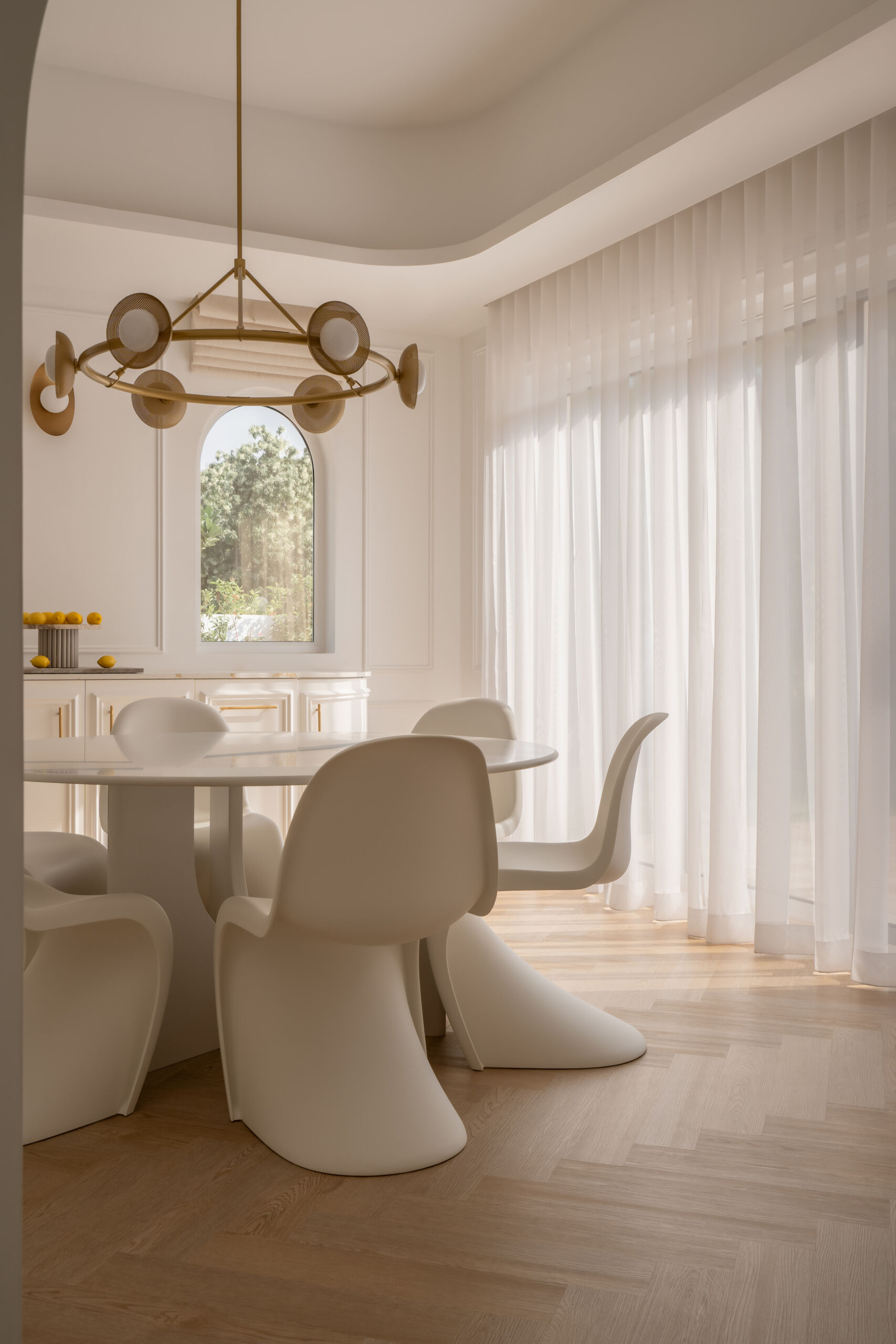 A contemporary casual dining room inside The White Arcade in Dubai, featuring sculptural white dining chairs, an oval table, a circular brass chandelier, arched windows, and elegant built-in cabinetry. Beautiful neutral dining room inspiration by D’Ora Tokai Designs.