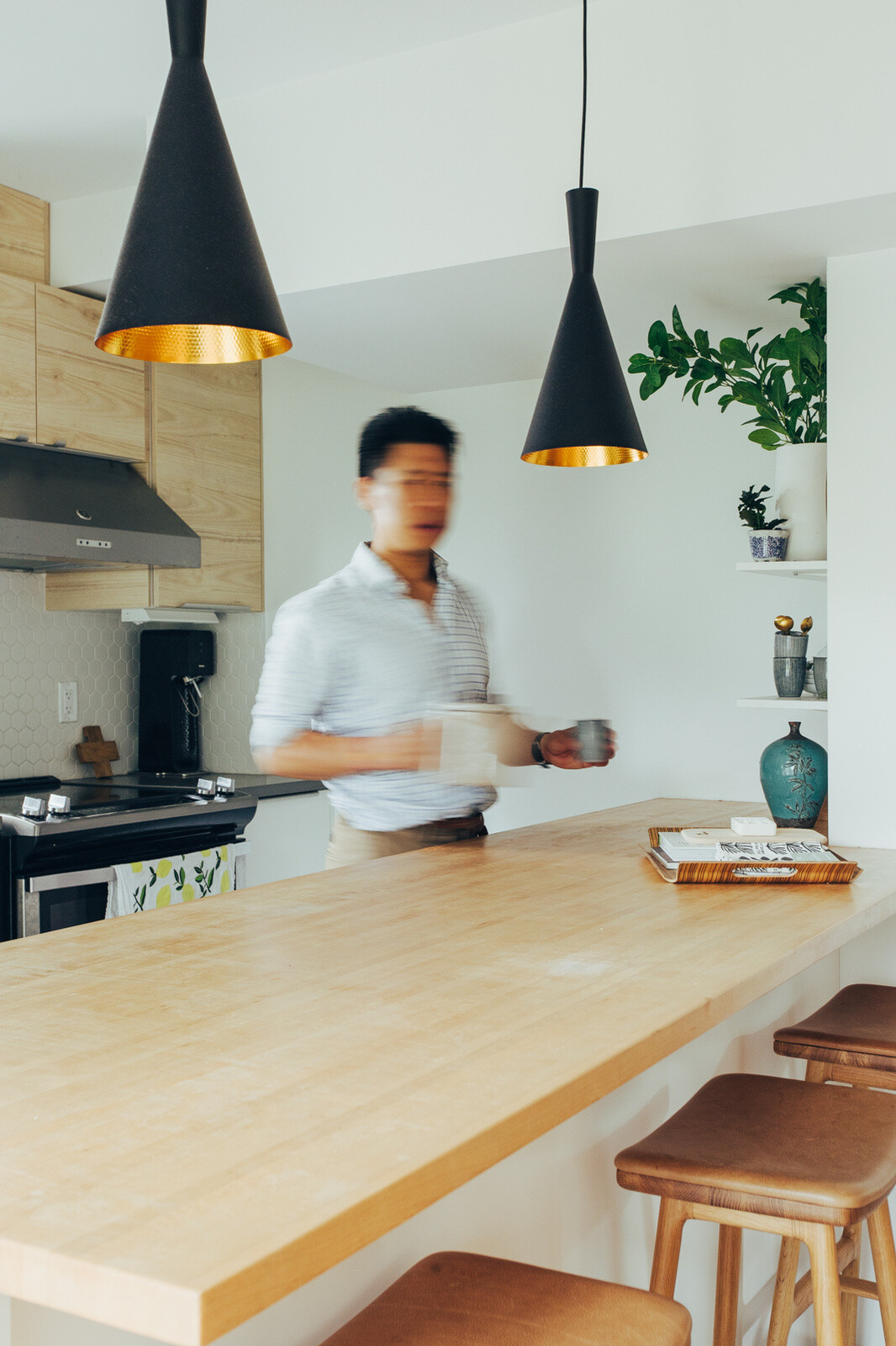This small contemporary kitchen features pale wood cabinets, a large wooden breakfast bar, black pendant lights, hexagonal backsplash tiles, and a timber slat partition.