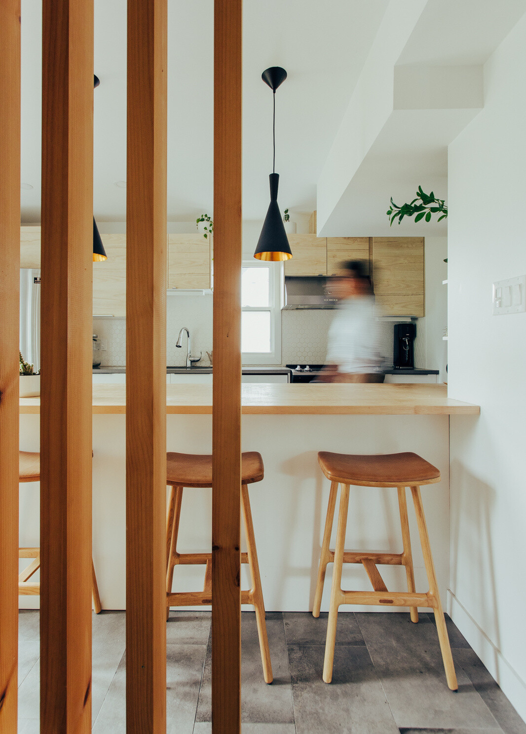 This small contemporary kitchen features pale wood cabinets, a large wooden breakfast bar, black pendant lights, hexagonal backsplash tiles, and a timber slat partition.