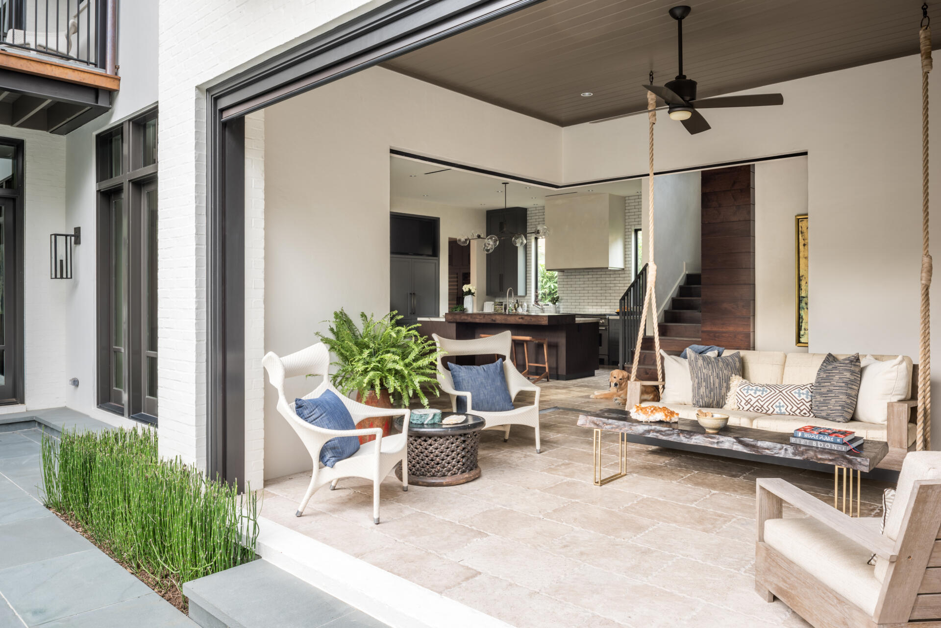 A covered outdoor lounge with a rope-suspended sofa, white woven chairs, blue cushions, rustic coffee table, and pocketing glass doors that open directly to the kitchen.