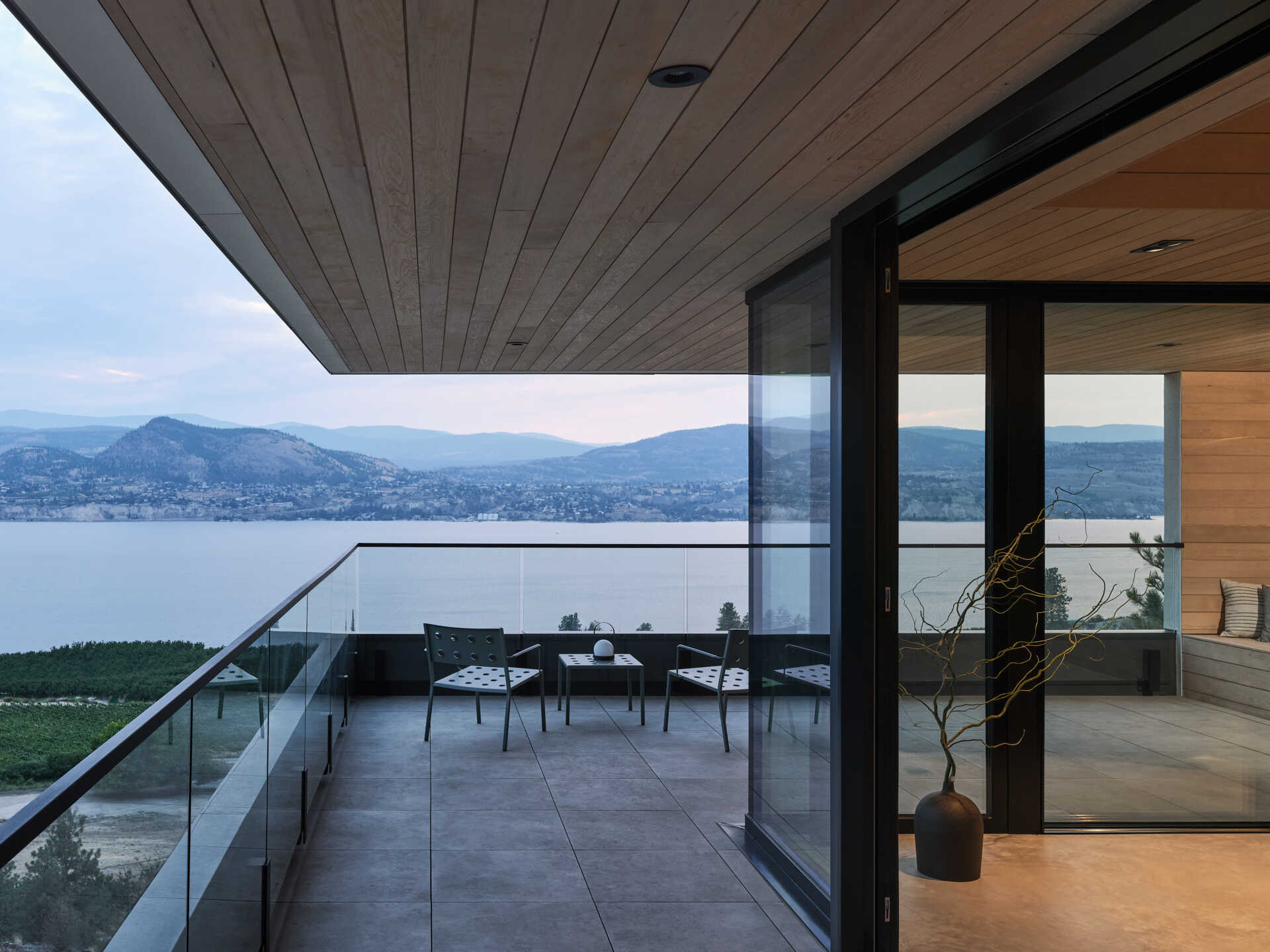 A covered terrace at a modern hillside house features gray floor tiles, black metal outdoor chairs, and glass railings overlooking Okanagan Lake. A deep wood-lined roof overhang provides shade while preserving the expansive view.