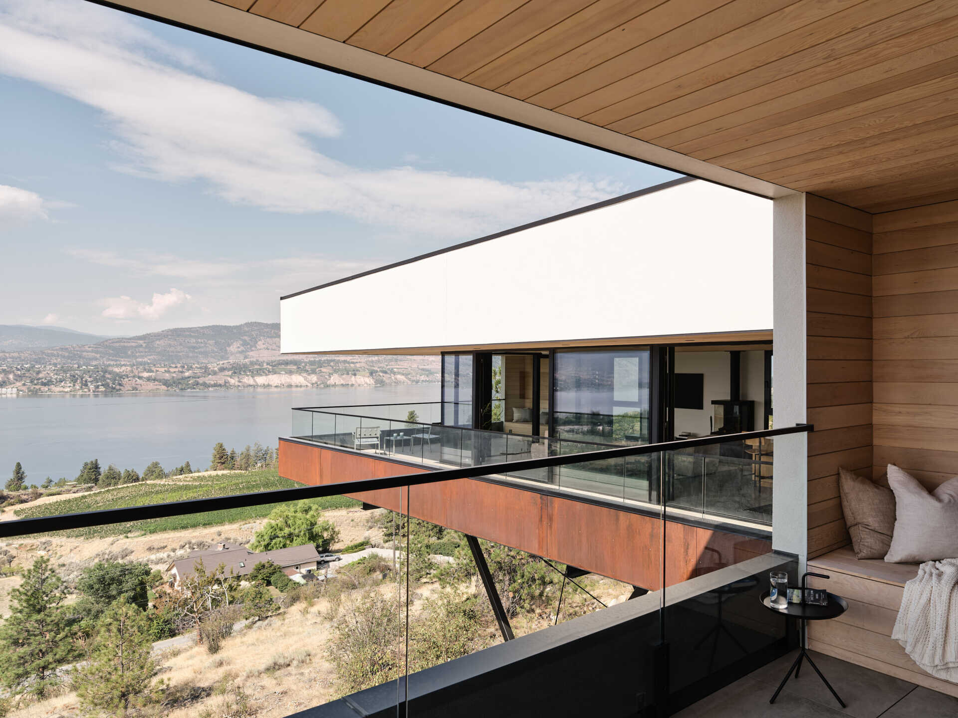 A covered terrace with wood-paneled walls, a built-in cushioned bench, and glass railings overlooks the cantilevered living wing of a modern hillside house. The projecting structure frames views toward Okanagan Lake.