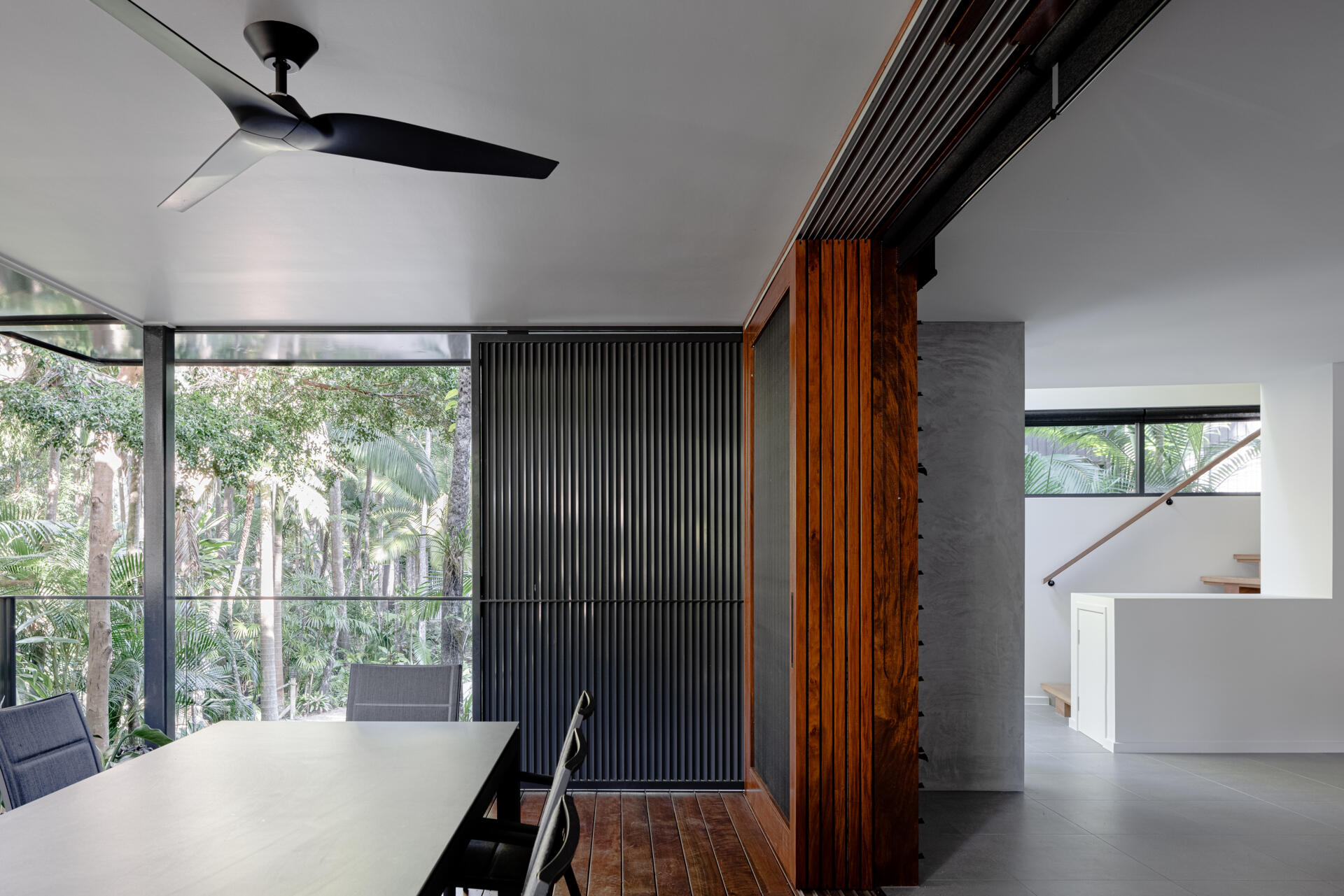 Explore the covered outdoor dining deck at Raintree in Noosa Heads, Australia. Designed by Void Building Design, the space features timber flooring, black vertical screens, a ceiling fan, and a dining table surrounded by tropical trees.