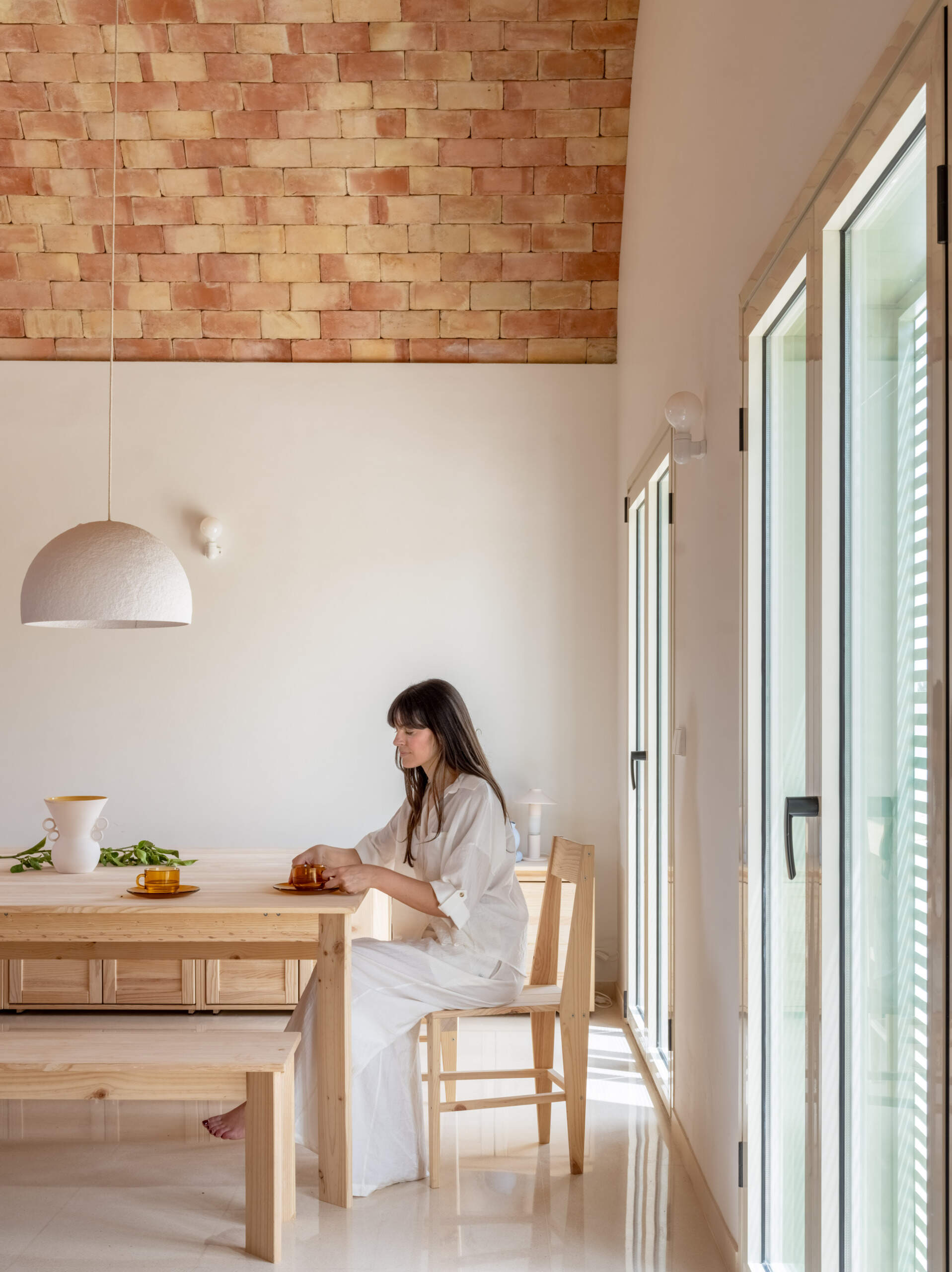 Natural Timber and Warm Cherry-Toned Cabinetry
Continuing through the open-plan interior, the dining area features a long, pale timber table paired with matching chairs and benches, creating a welcoming space for shared meals. A simple white dome pendant hangs above the table, while a low timber sideboard runs along the adjacent wall. The exposed brick ceiling stretches overhead, bringing warmth and texture to the otherwise minimal space. Large glass doors allow sunlight to filter through the shutters, filling the room with natural light.
Beside the dining area, the kitchen introduces a richer material palette through cabinetry made from pine lacquered in a cherry tone. The reddish timber contrasts with the light-coloured countertops, while a long kitchen island provides additional workspace and casual seating with timber stools. Integrated appliances, streamlined cabinetry, and minimal detailing keep the kitchen looking uncluttered, allowing the natural materials and vaulted ceiling to remain the main features.