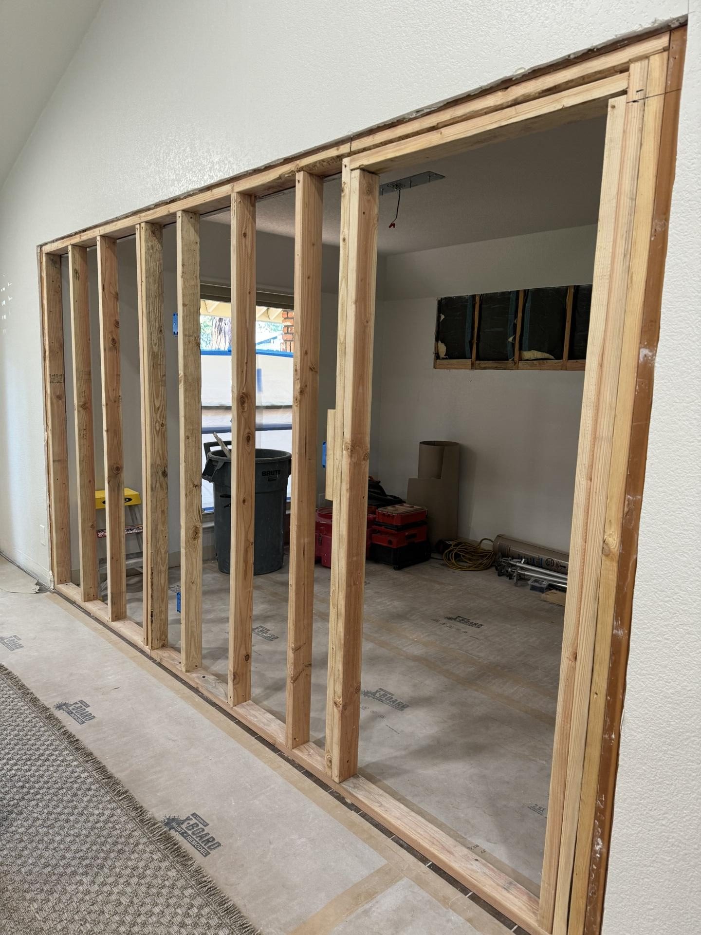 Wood wall framing takes shape during the conversion of an open dining room into an enclosed home office. The construction photograph shows the new wall studs, exposed interior, and protected flooring.