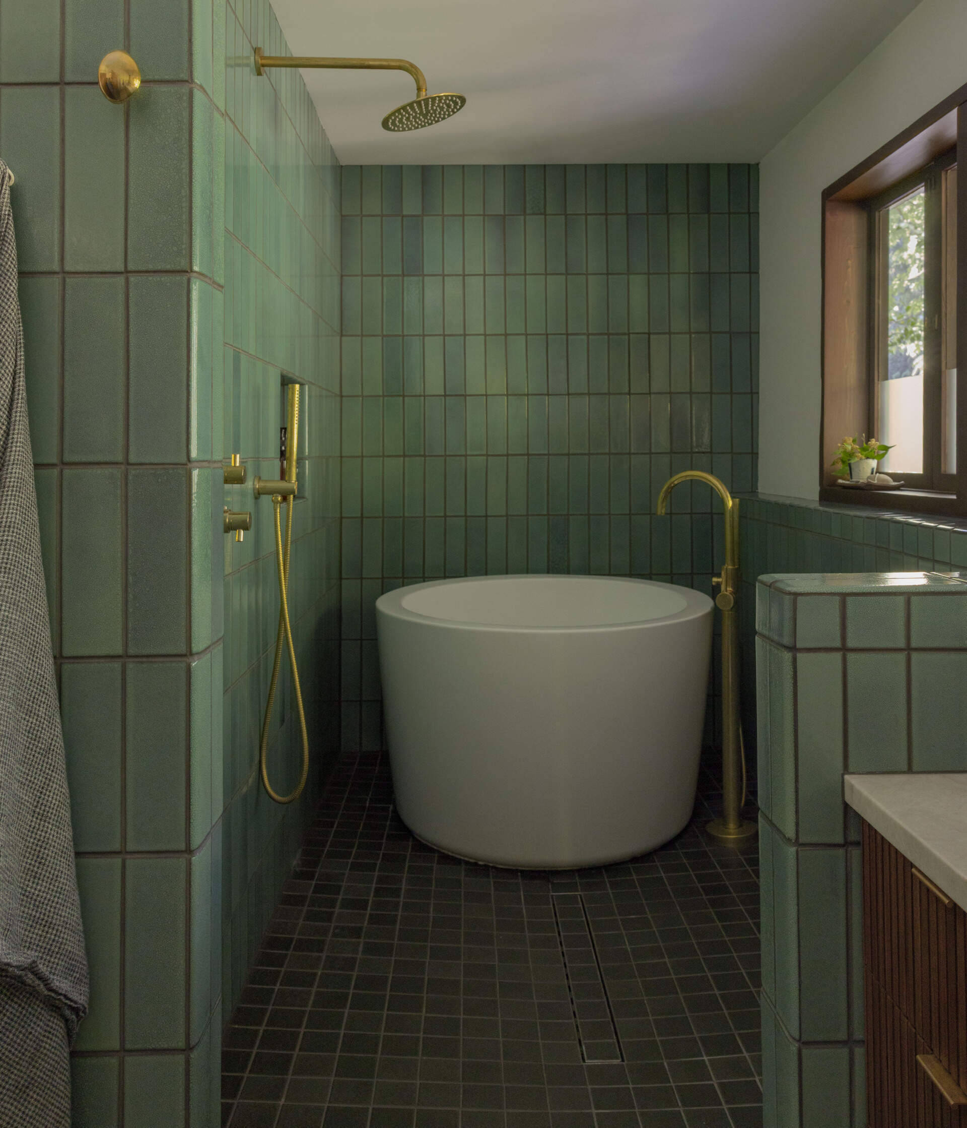 Explore the primary ensuite bathroom of The Burrows, a renovated mid-century home in Seattle by chadbourne + doss architects. Green Heath tiles, a freestanding soaking tub, brass fixtures, and a custom walnut double vanity create a beautiful modern bathroom.