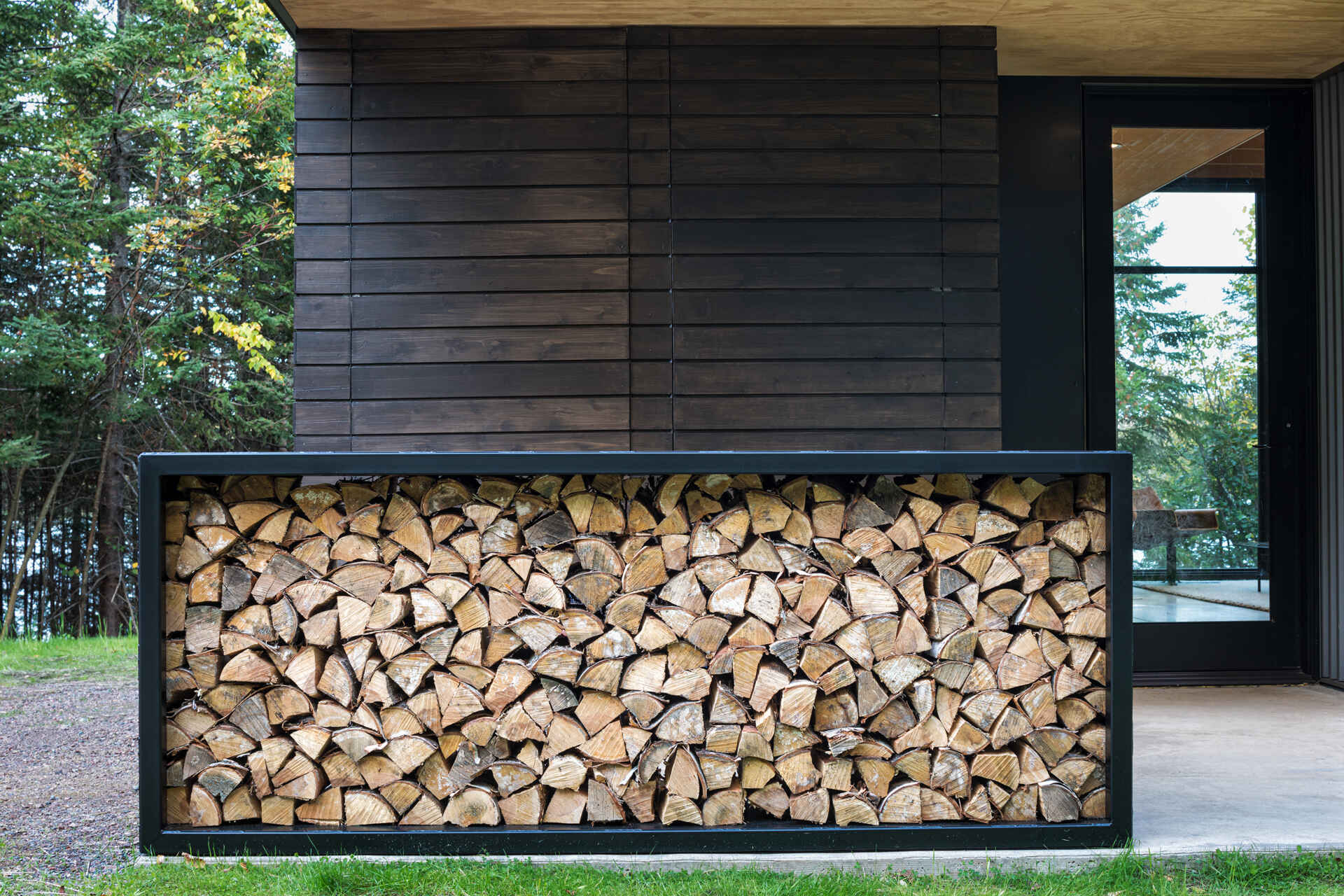 A built-in steel firewood storage box filled with neatly stacked logs beside the entrance of a modern cabin. Dark horizontal cedar siding and a glass entrance door complete the exterior detail.