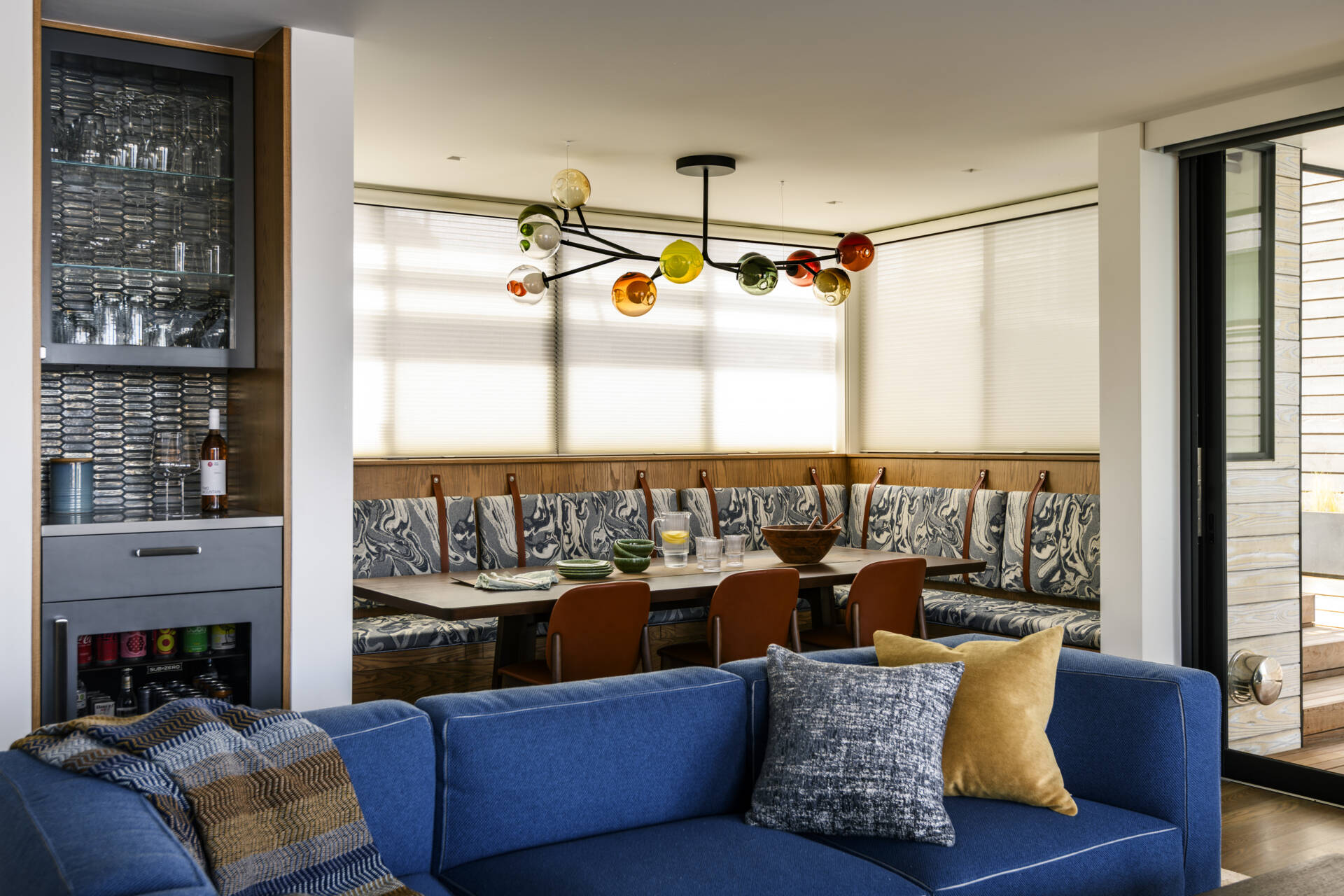 An L-shaped upholstered banquette, warm wood dining table, cognac-colored chairs, and sculptural multicolored glass chandelier create a fun and welcoming dining space.