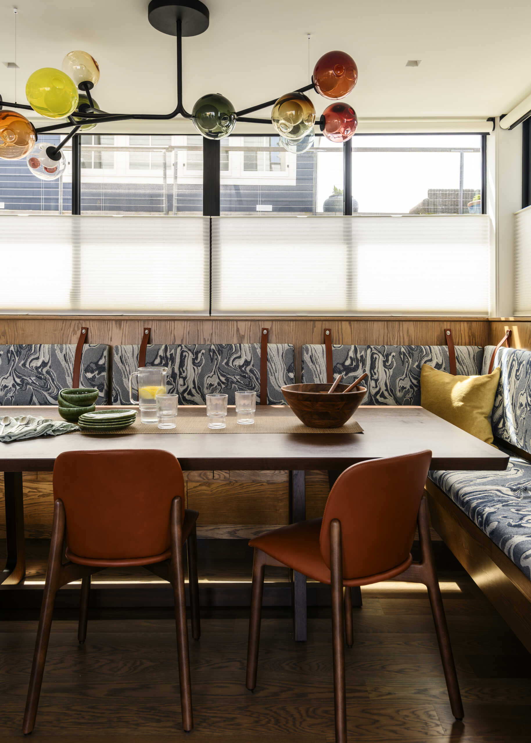 An L-shaped upholstered banquette, warm wood dining table, cognac-colored chairs, and sculptural multicolored glass chandelier create a fun and welcoming dining space.