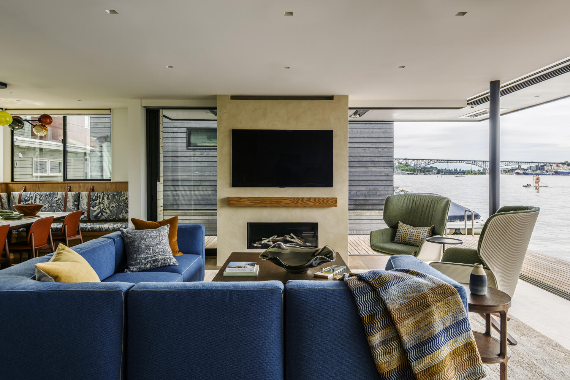 Step inside the living room of Modern Beach House, a floating home on Lake Union in Seattle by Hoshide Wanzer Architects + Interiors. A blue sectional sofa, green lounge chairs, double-sided fireplace, and large pocketing glass doors create a contemporary waterfront living space with uninterrupted lake views.