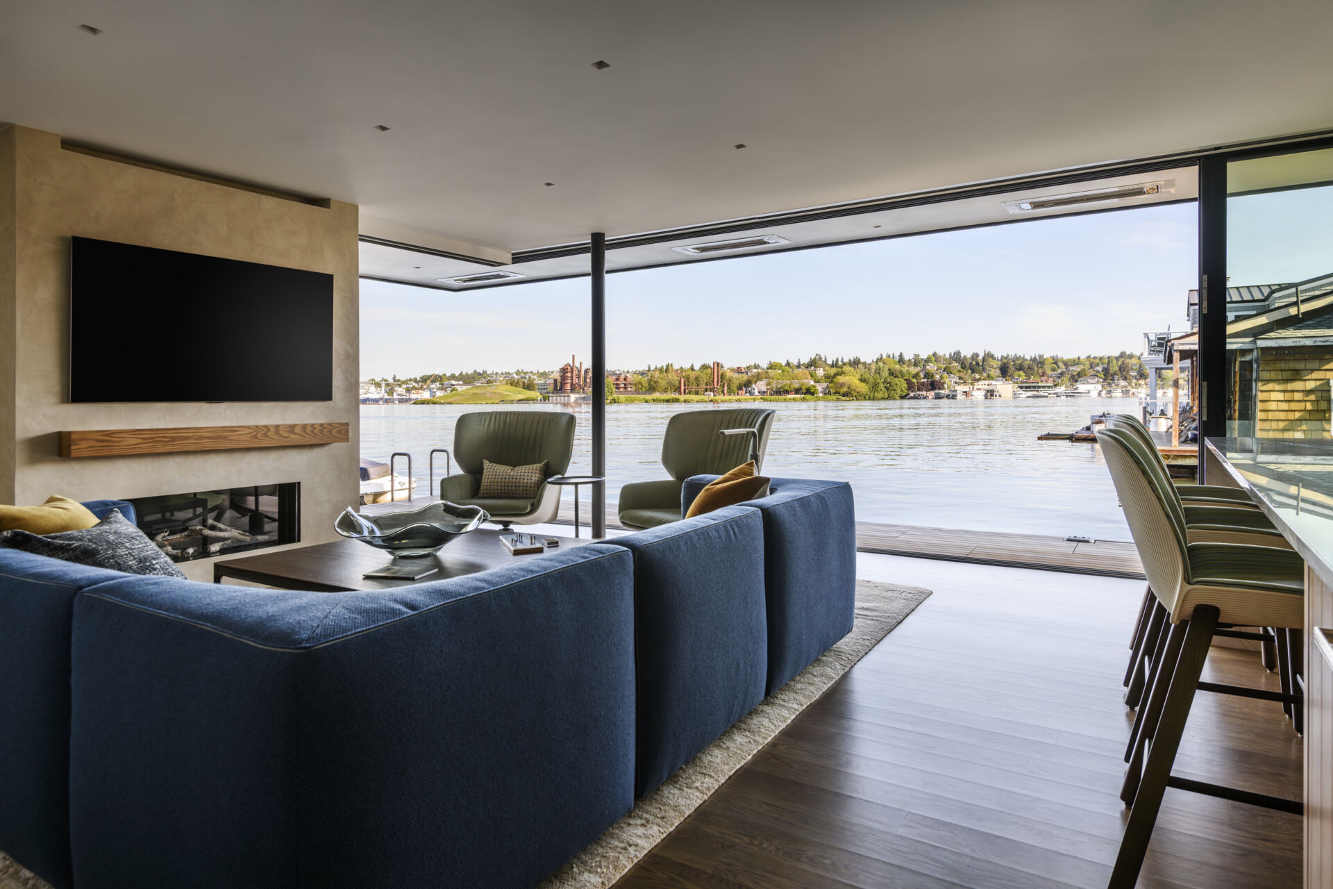 Step inside the living room of Modern Beach House, a floating home on Lake Union in Seattle by Hoshide Wanzer Architects + Interiors. A blue sectional sofa, green lounge chairs, double-sided fireplace, and large pocketing glass doors create a contemporary waterfront living space with uninterrupted lake views.