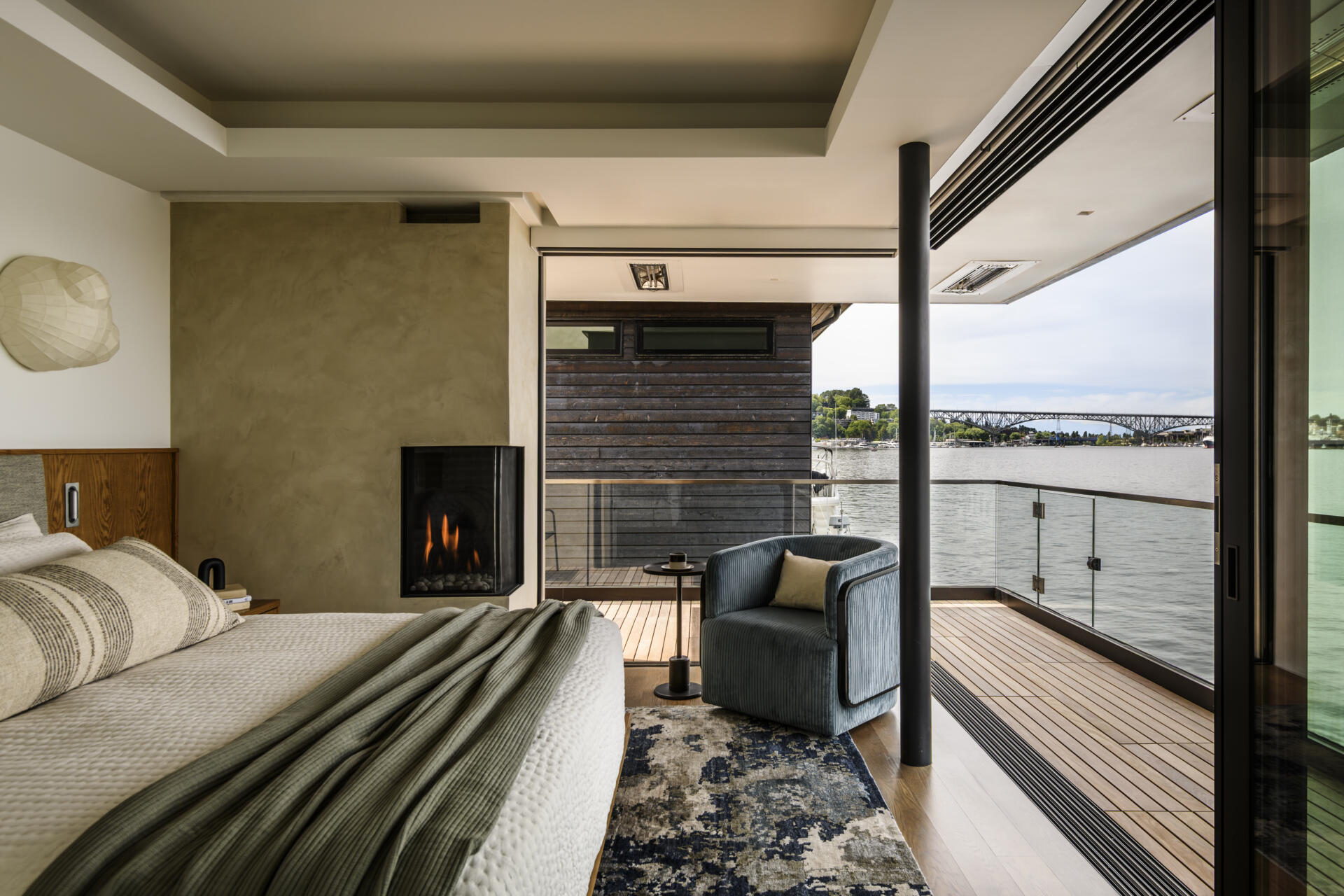 Featuring a private waterfront balcony, corner fireplace, upholstered headboard, built-in wood cabinetry, and soft coastal colors, this modern bedroom offers beautiful lake views.