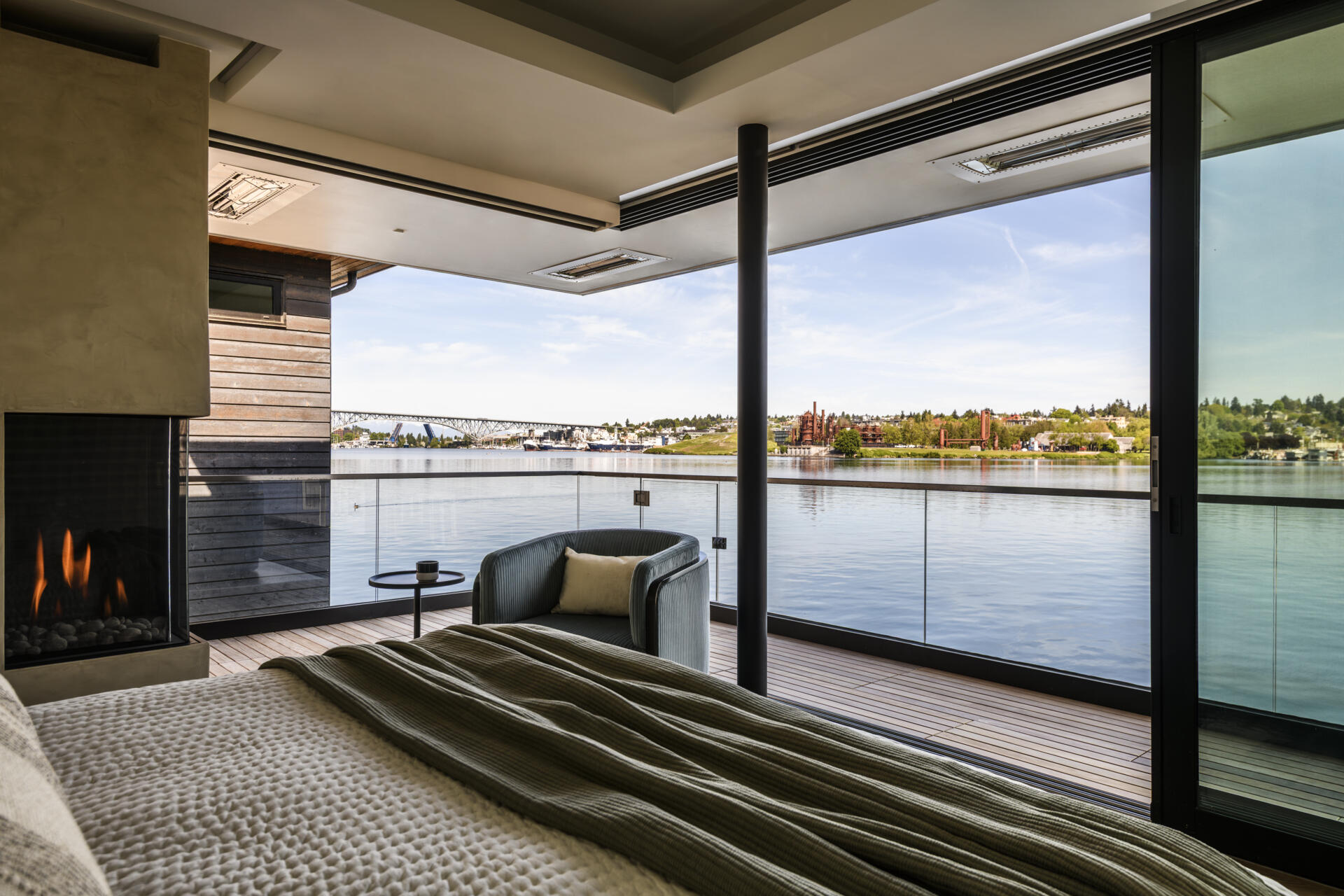 Featuring a private waterfront balcony, corner fireplace, upholstered headboard, built-in wood cabinetry, and soft coastal colors, this modern bedroom offers beautiful lake views.