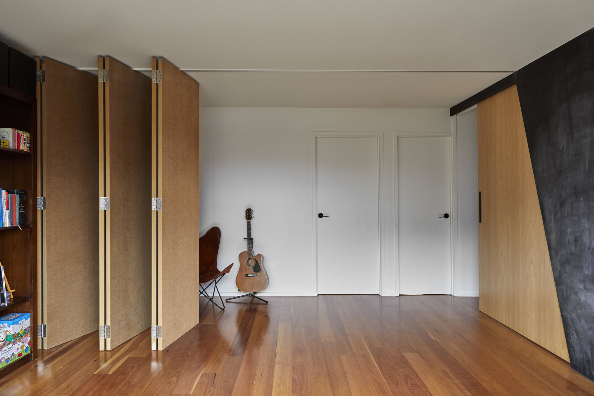 Full-height folding wall panels are stacked against one side of a room, revealing timber floors, white walls, and a seating area with a leather chair and guitar.