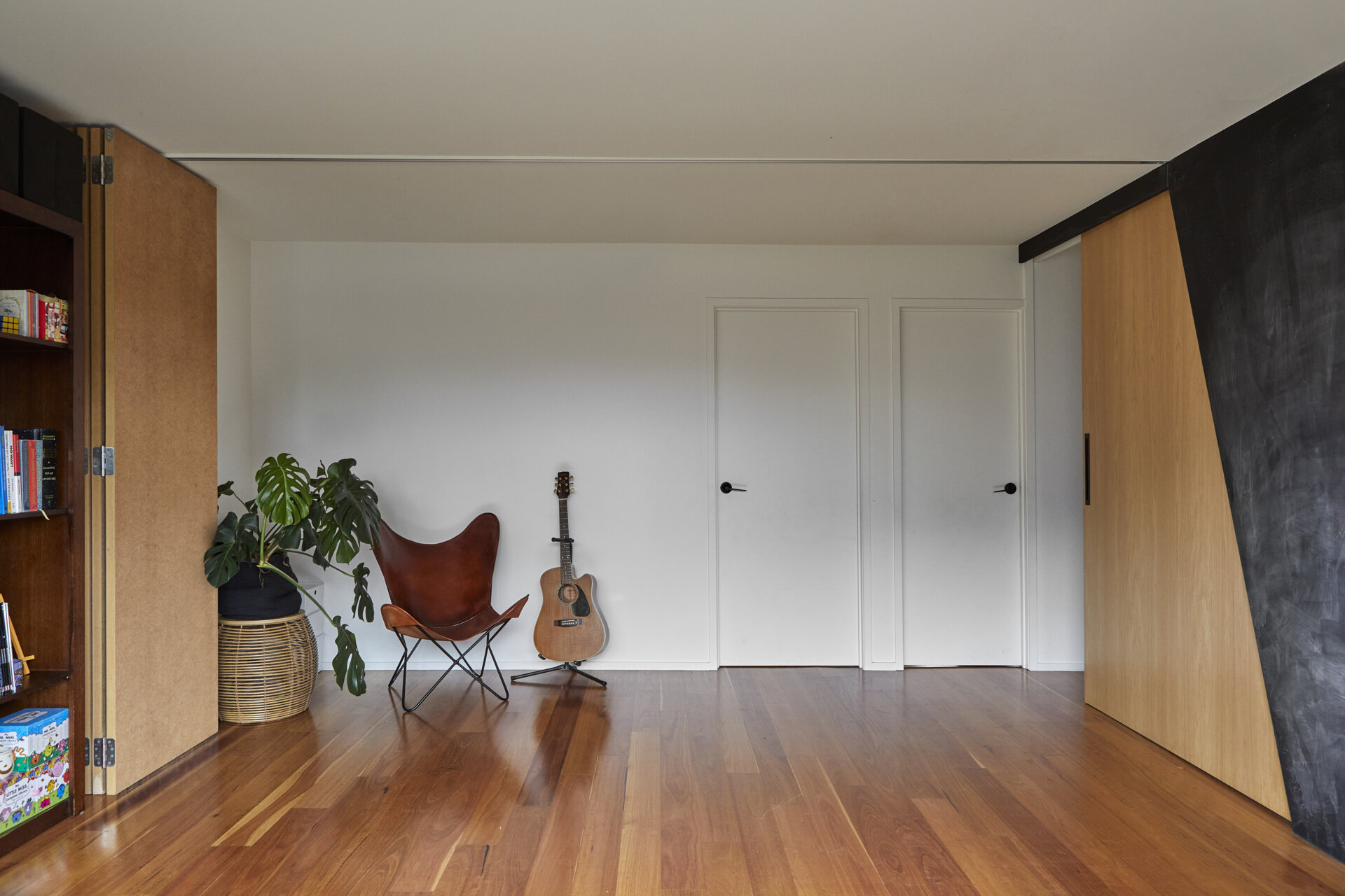 A folding partition opens up a modern family room with timber flooring, white walls, and a small seating area. The panels fold away beside a built-in bookcase.