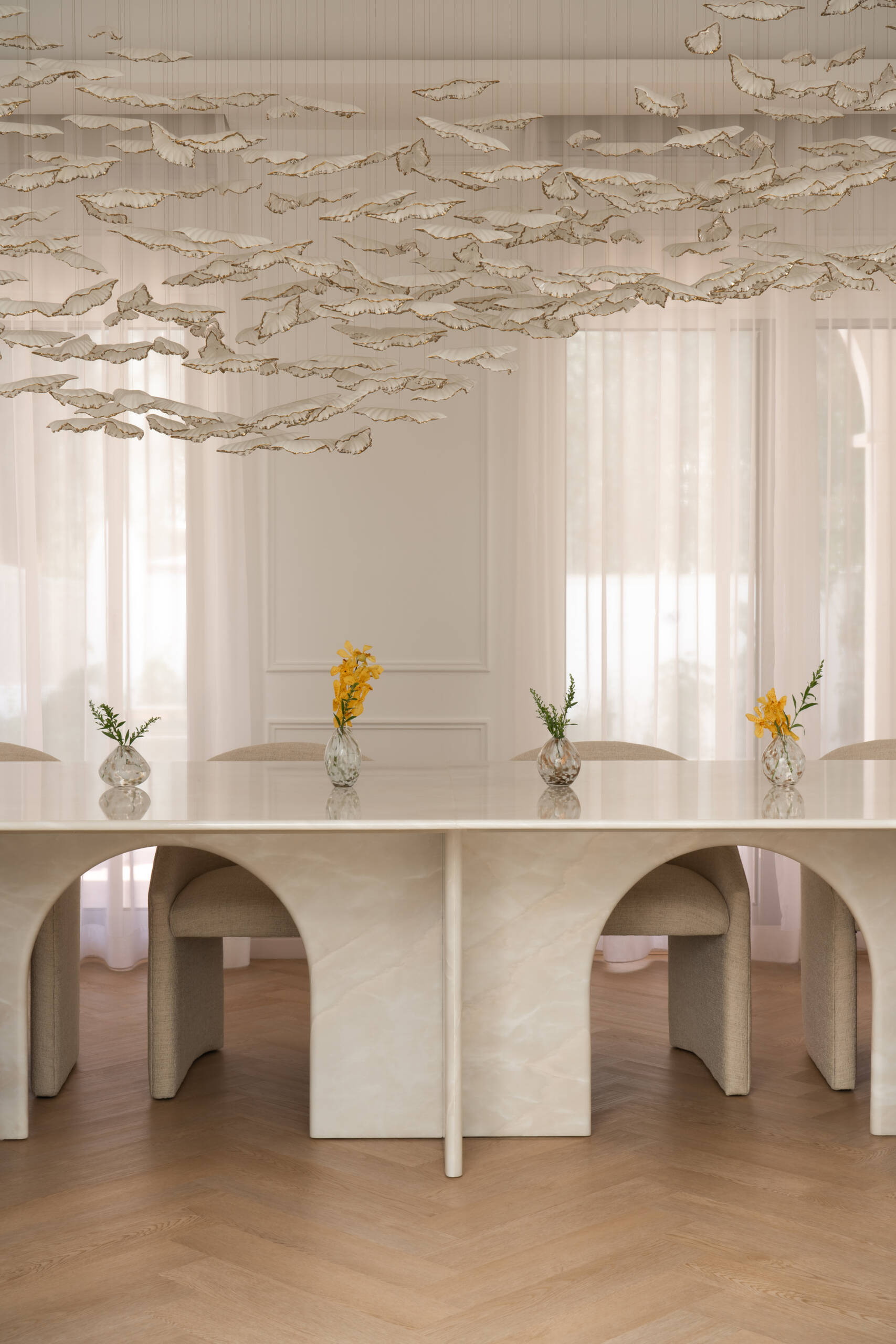 This luxurious formal dining room in Dubai features a monumental white onyx dining table by DOT Objects, sculptural arched bases, curved upholstered dining chairs, and an incredible suspended leaf installation. Designed by D’Ora Tokai Designs for The White Arcade.