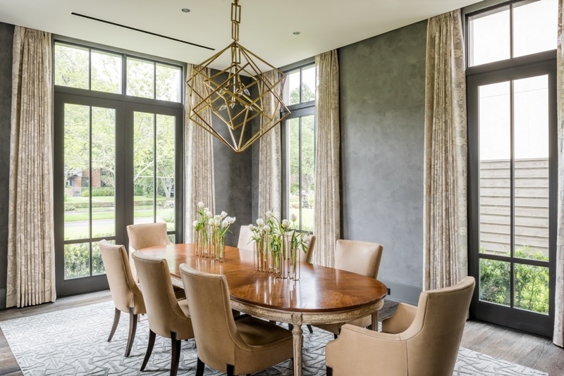 A formal dining room featuring charcoal-gray textured walls, an oval wood dining table, cream upholstered chairs, colorful abstract art, and a sculptural gold geometric chandelier.